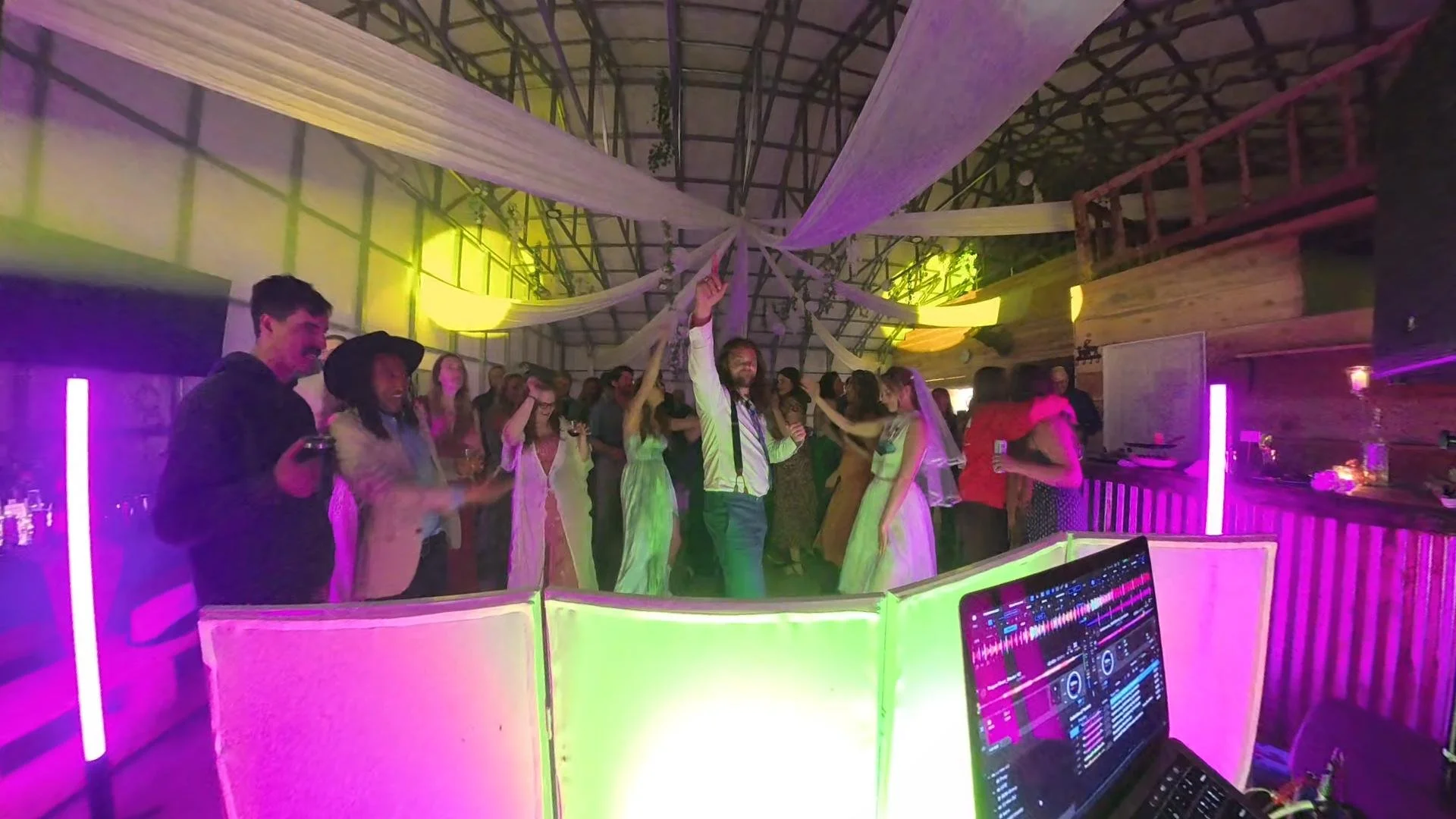 People dancing and socializing in a party with colorful neon lights, a DJ booth with a computer, and a wooden interior ceiling with drapes hanging down.