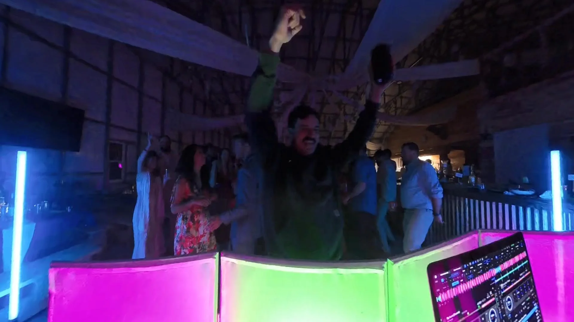 Man dancing with his arms raised inside a nightclub with red and blue neon lighting, surrounded by people. DJ equipment visible in the foreground.