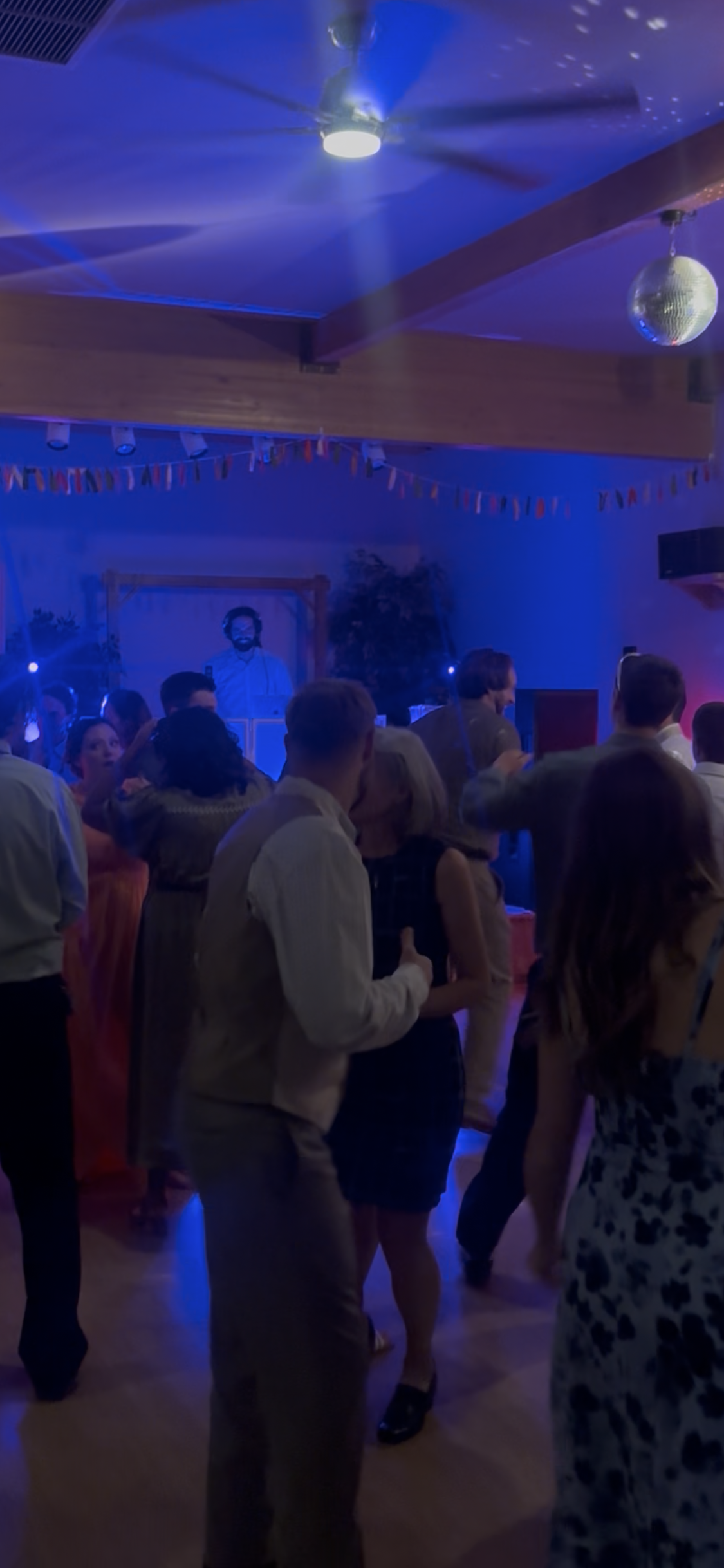 People dancing at a party with a DJ playing music in the background. There is a disco ball hanging from the ceiling and colorful party lights.