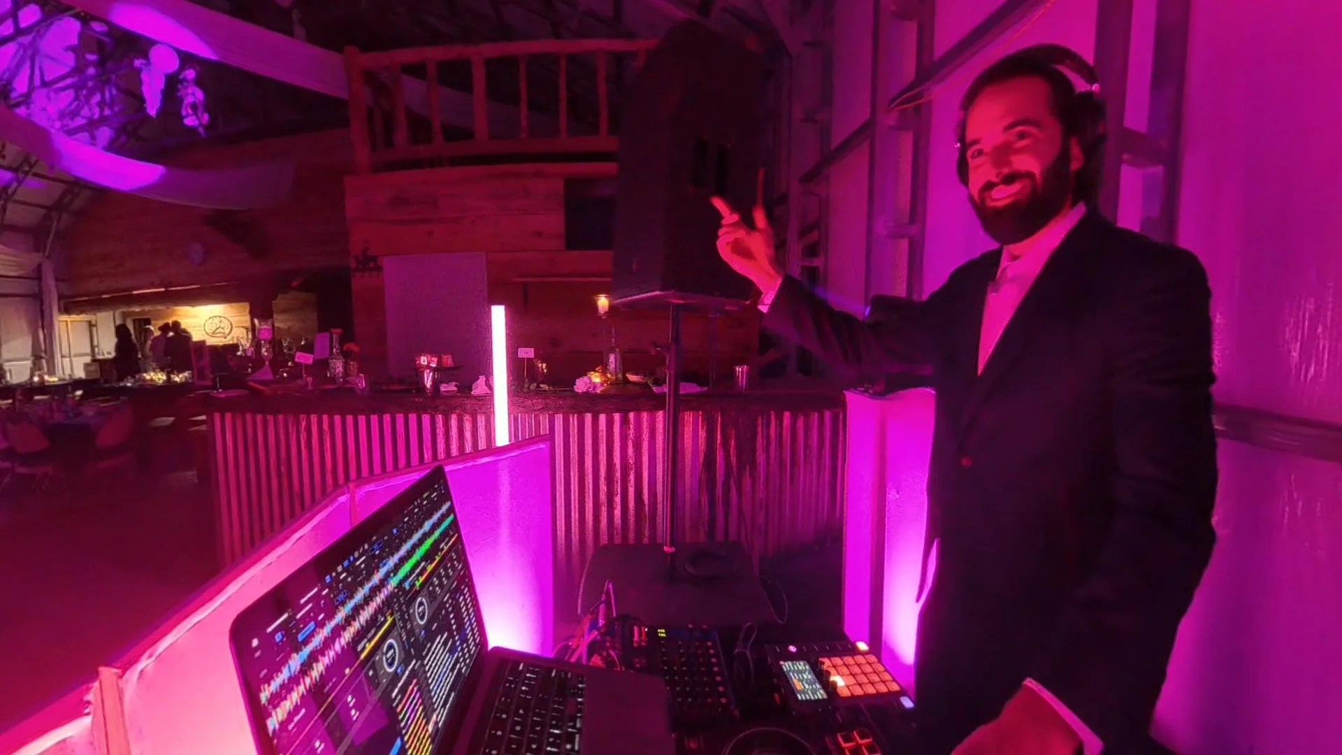 A DJ performing at a party with purple lighting, wearing a suit and smiling at the camera, with DJ equipment and a laptop in front.