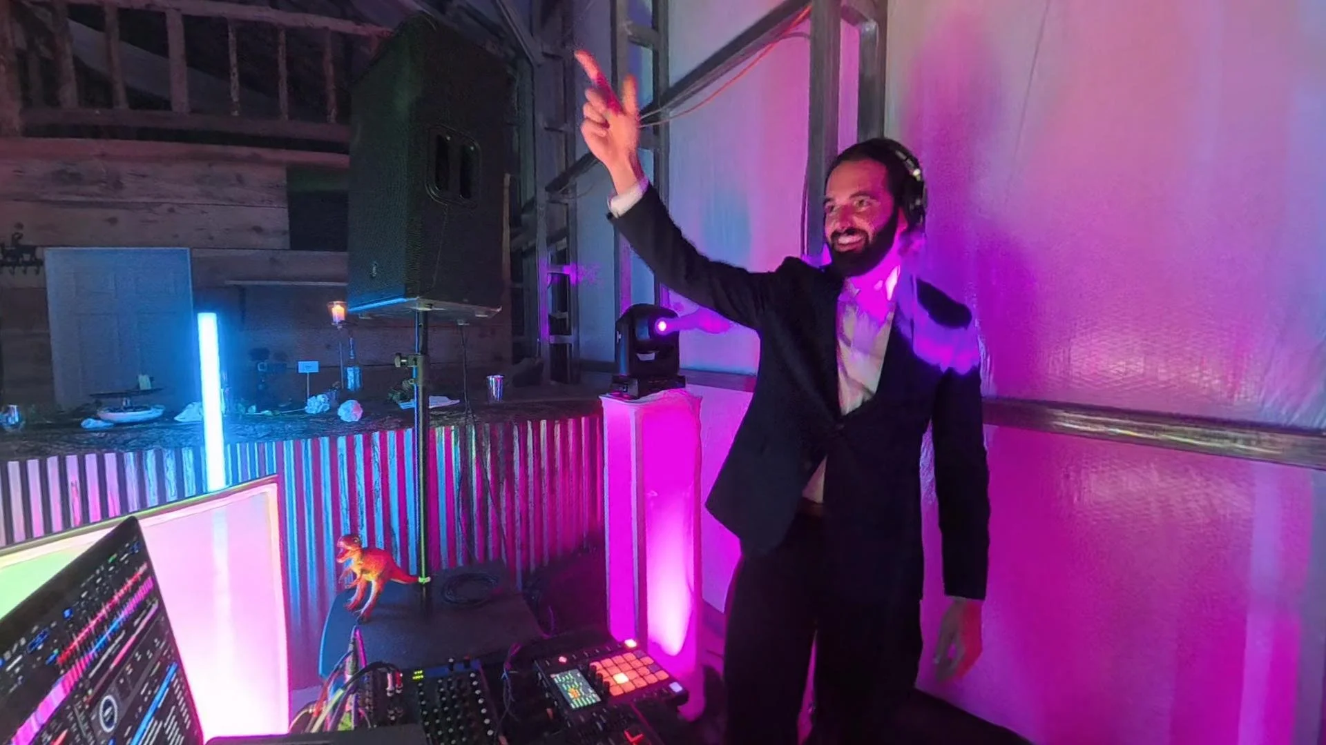 A man in a suit and headphones smiling and dancing at a DJ booth with colorful lighting, DJ equipment, and a decorated table in the background.