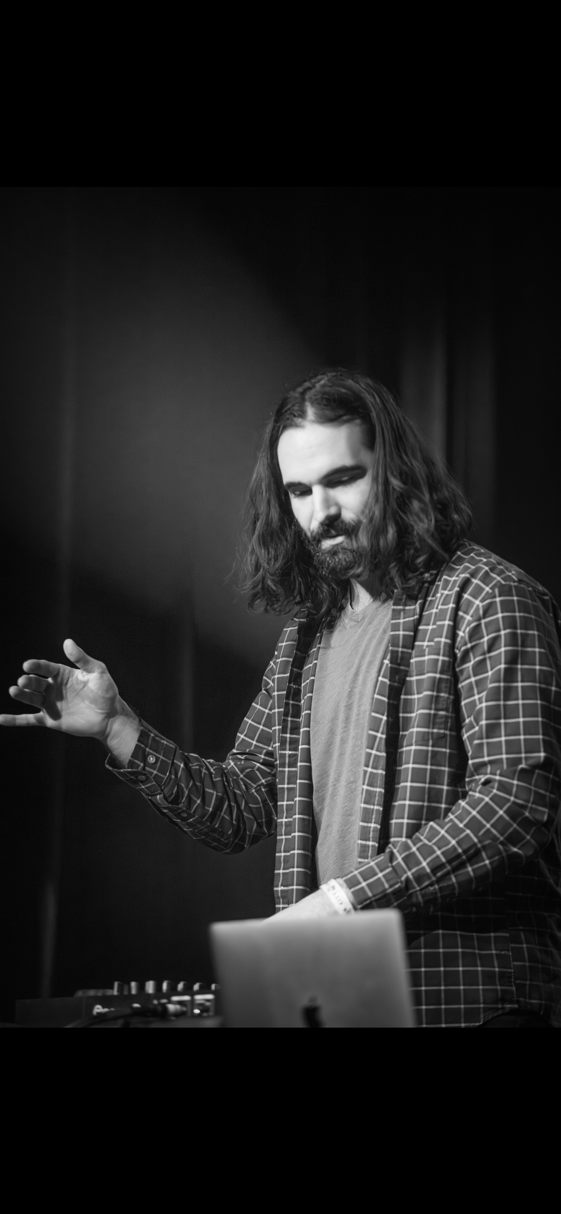 Black and white photo of a man with long hair and a beard, wearing a checkered shirt and a T-shirt, standing in front of DJ equipment, with his eyes closed and hand raised.