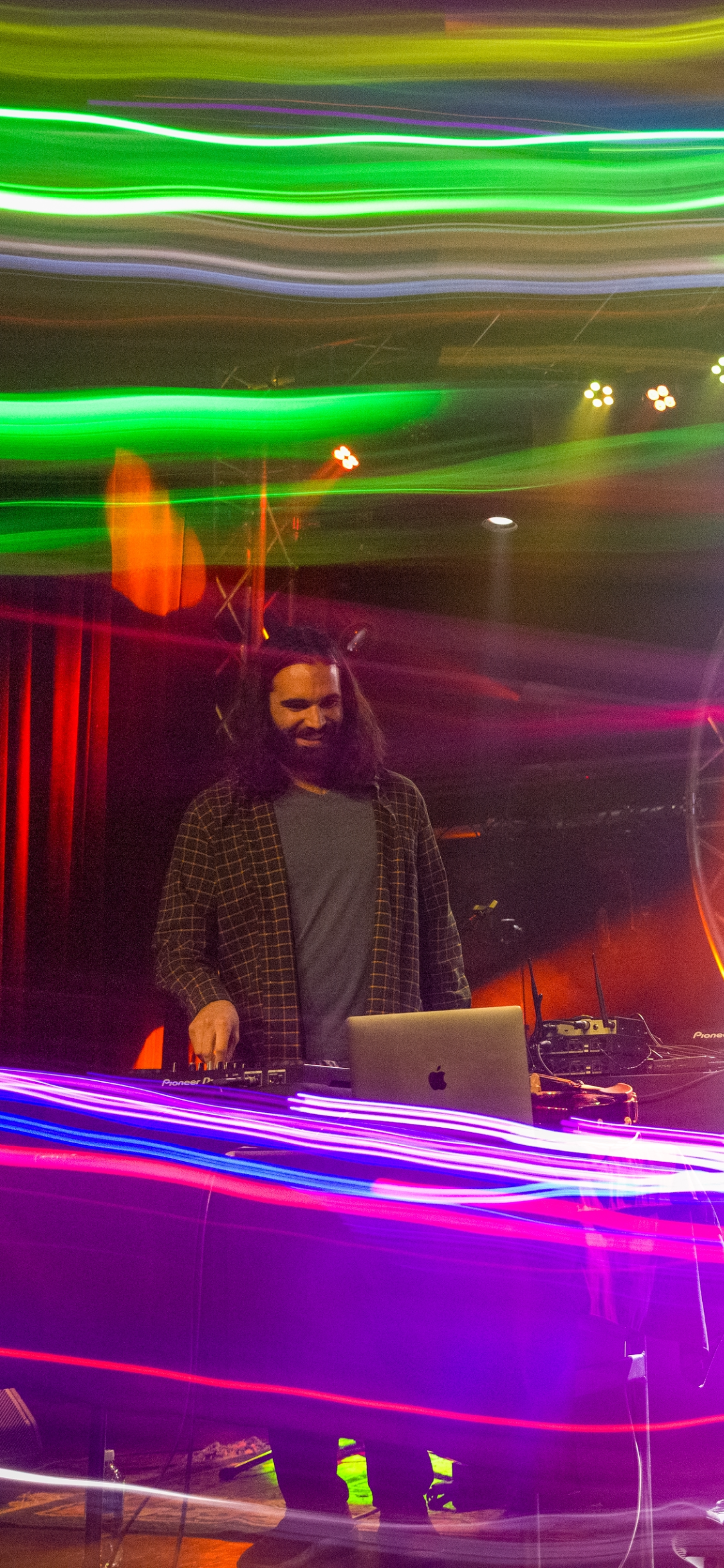 DJ performing at a club with colorful light streaks in the background, smiling while operating DJ equipment and a laptop.