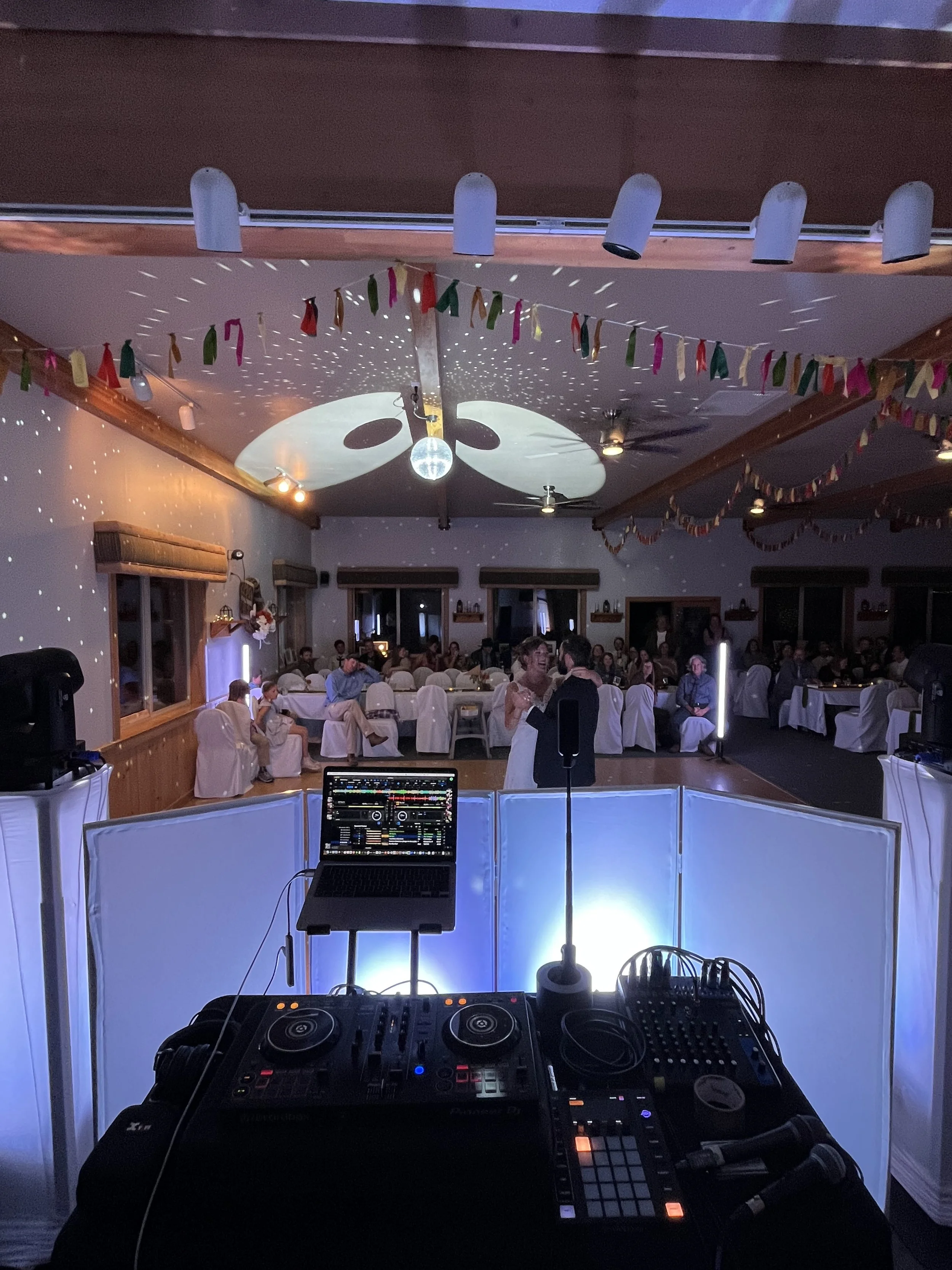 Indoor party or event with people sitting at decorated tables, colorful bunting, a disco ball, and a DJ setup with lighting effects.