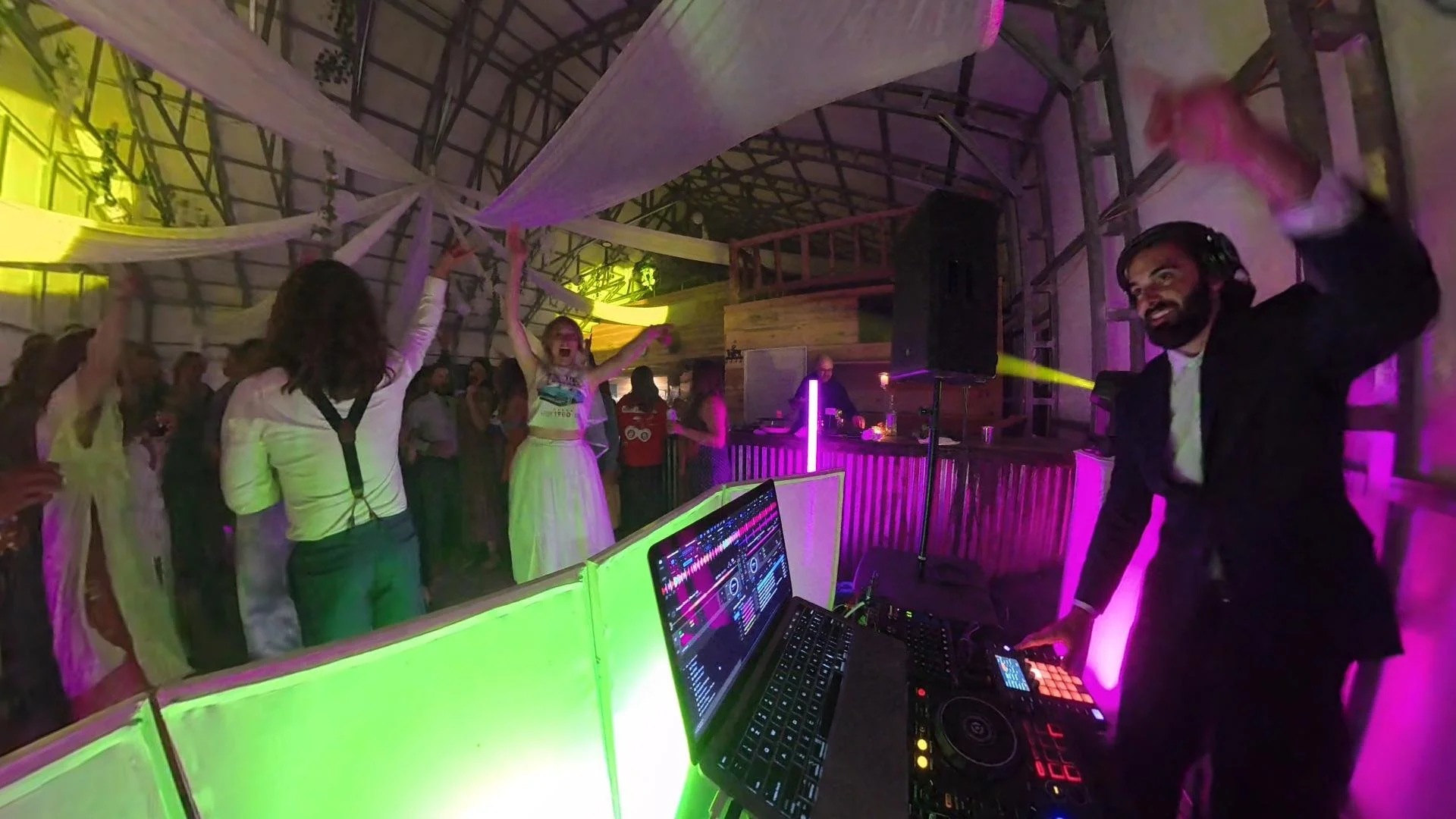 People dancing at a lively party with colorful lighting, a DJ setup in the foreground, and a wooden structure interior.
