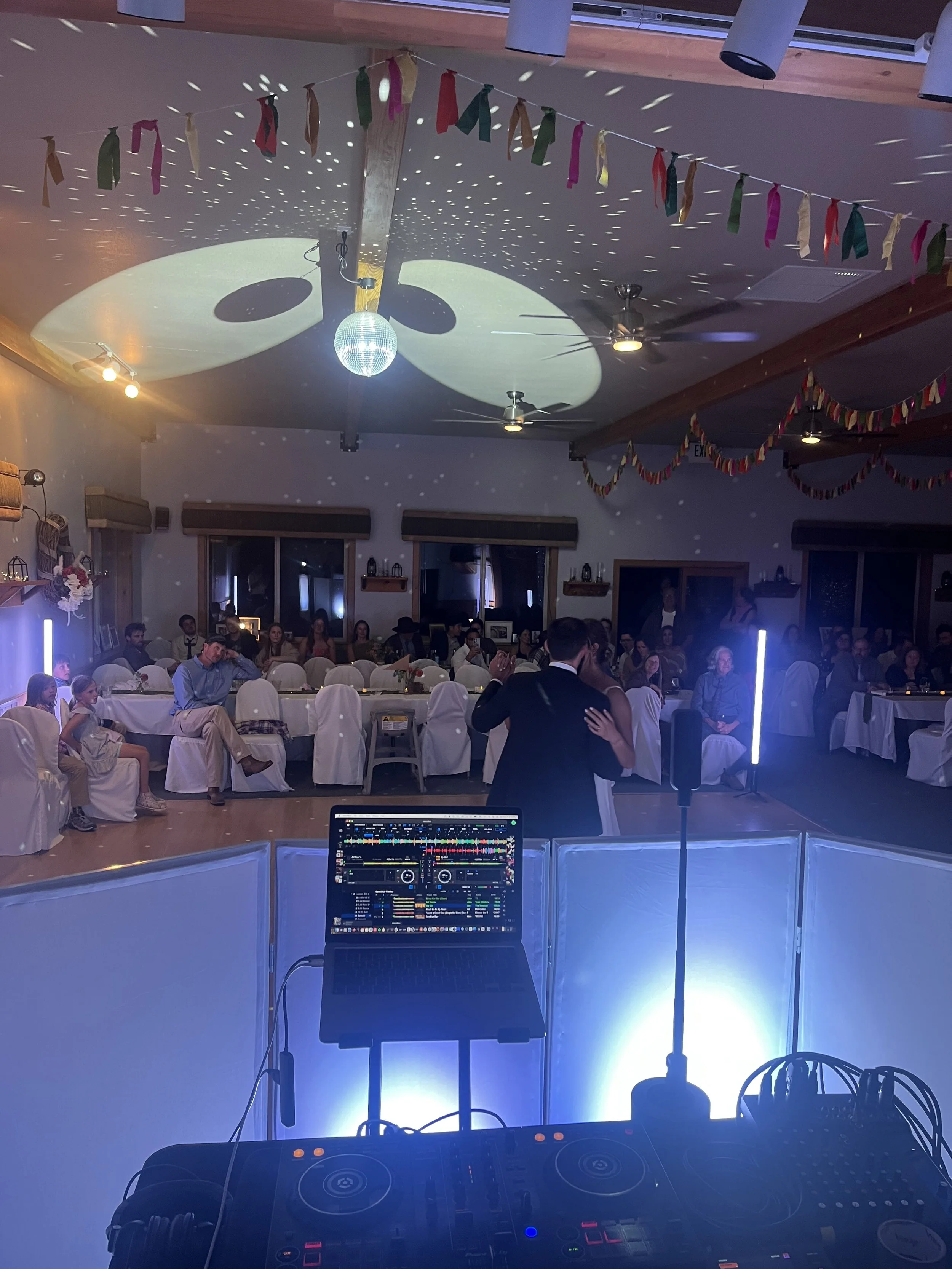 A large room decorated with colorful flags and garlands, filled with people sitting at tables, with a DJ setup in the foreground, and a disco ball casting lights on the ceiling.