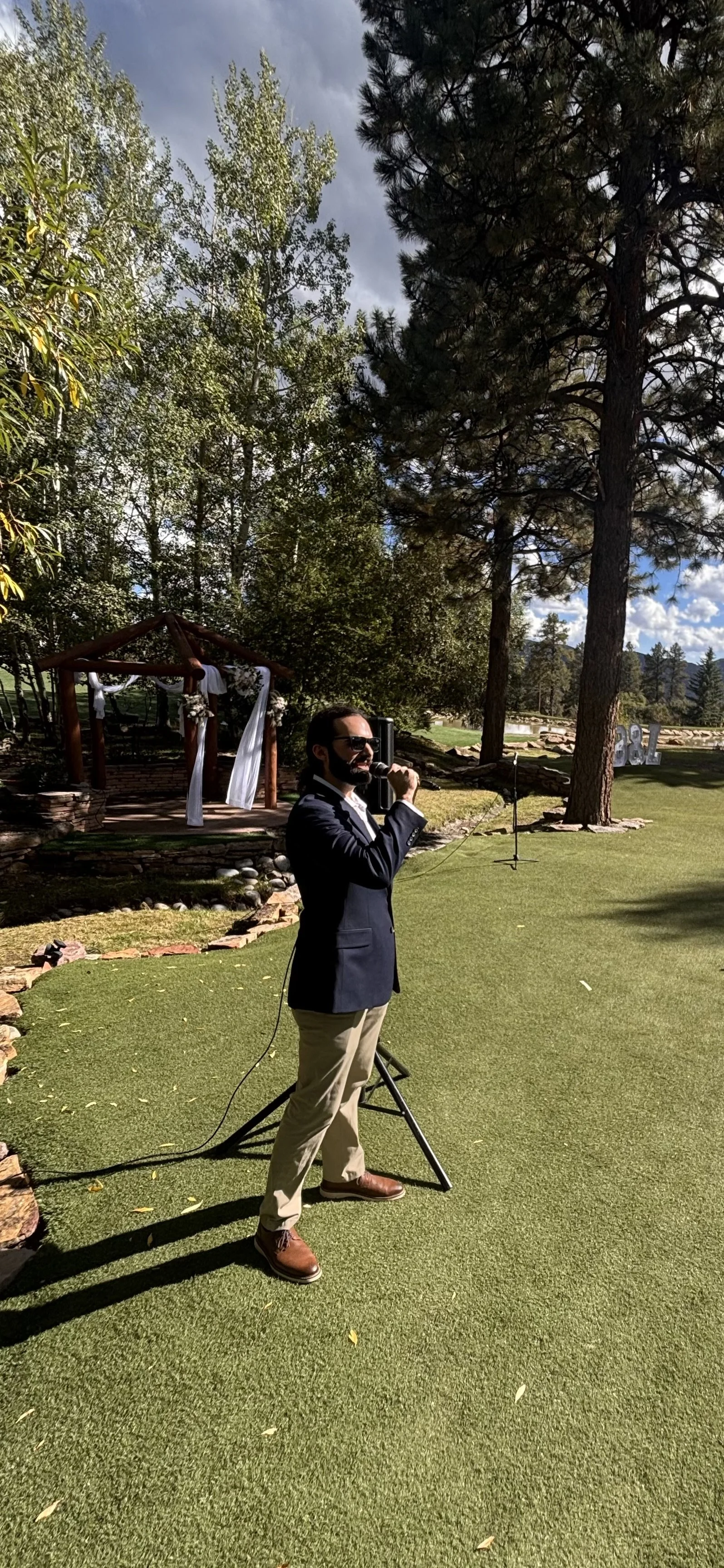 Man in a suit and beige pants holding a microphone and speaking outdoors in front of a green lawn and trees with cloudy sky.