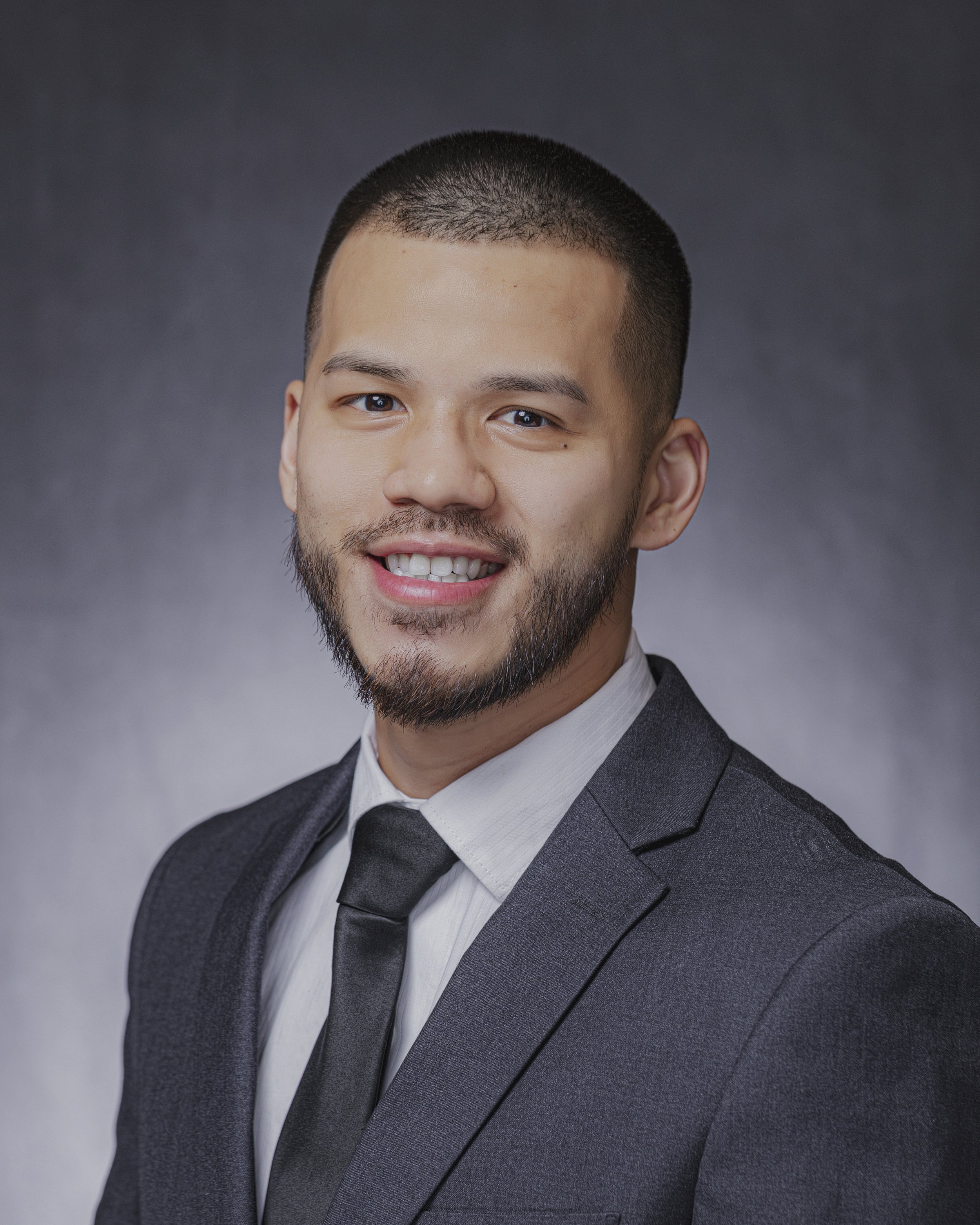 Portrait of a young Asian man with short black hair, a well-groomed beard, wearing a dark gray suit, white shirt, and black tie, smiling against a neutral gray background.