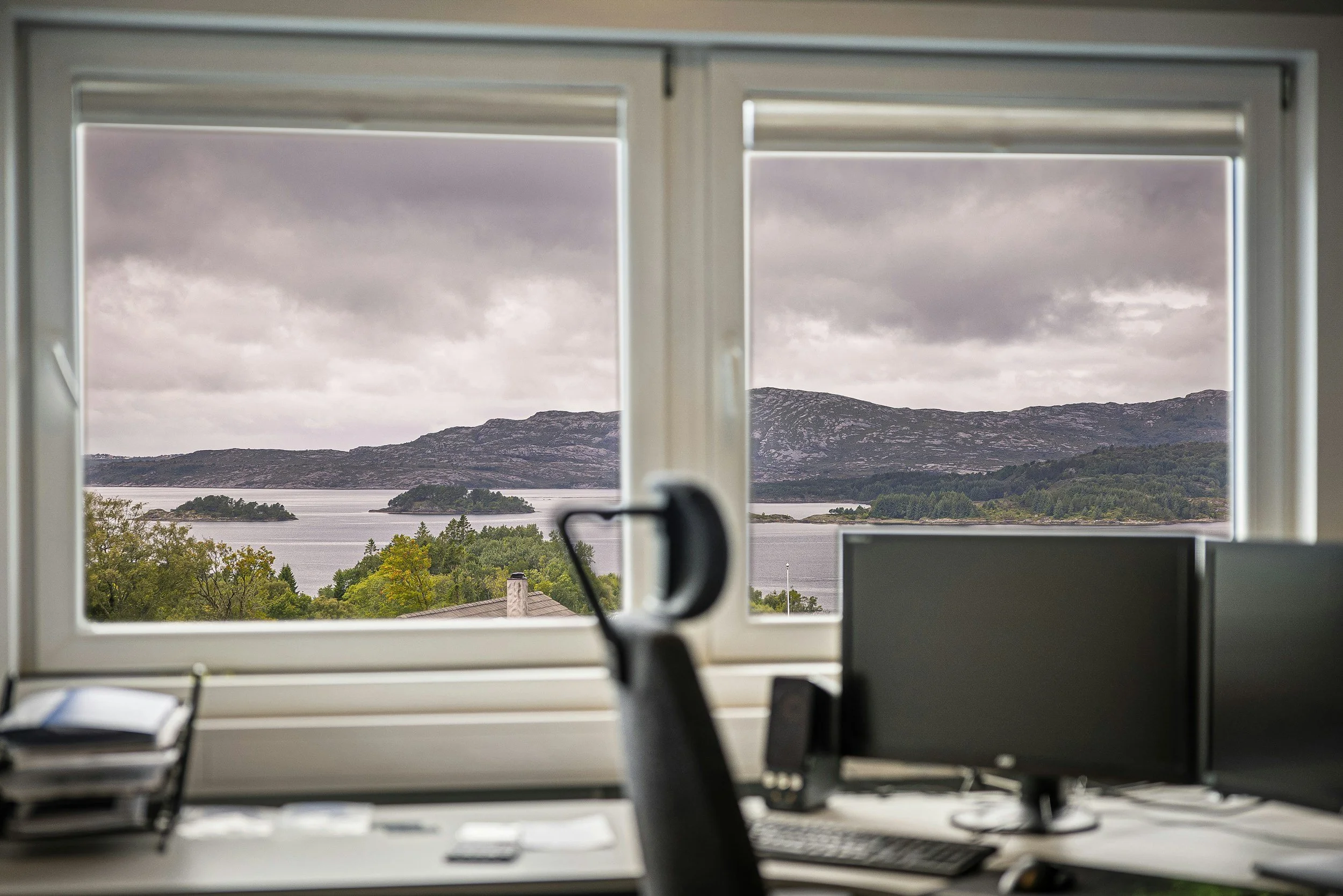 View of a mountains and water landscape through a window from an indoor office or room, with office desks and computer monitors visible in the foreground.