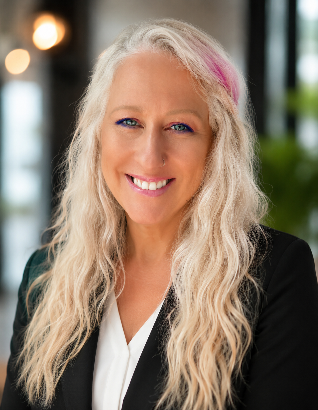 A smiling woman with long blonde wavy hair, wearing a black blazer and white blouse, in an indoor setting with blurred background.
