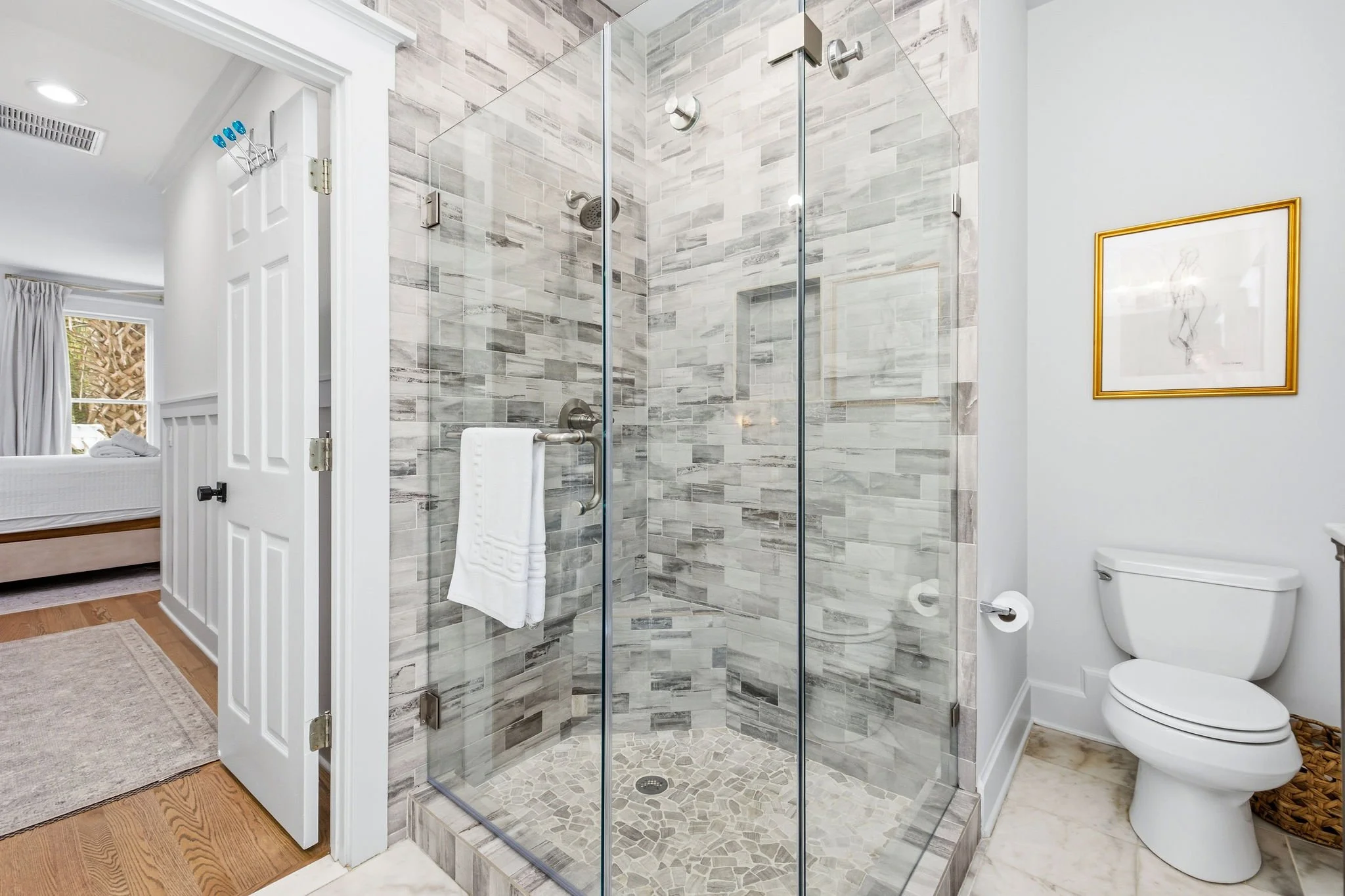 Bathroom with a glass shower enclosure, beige and gray tiled walls, a white toilet, and framed artwork on the wall. Part of a bedroom is visible through an open door.