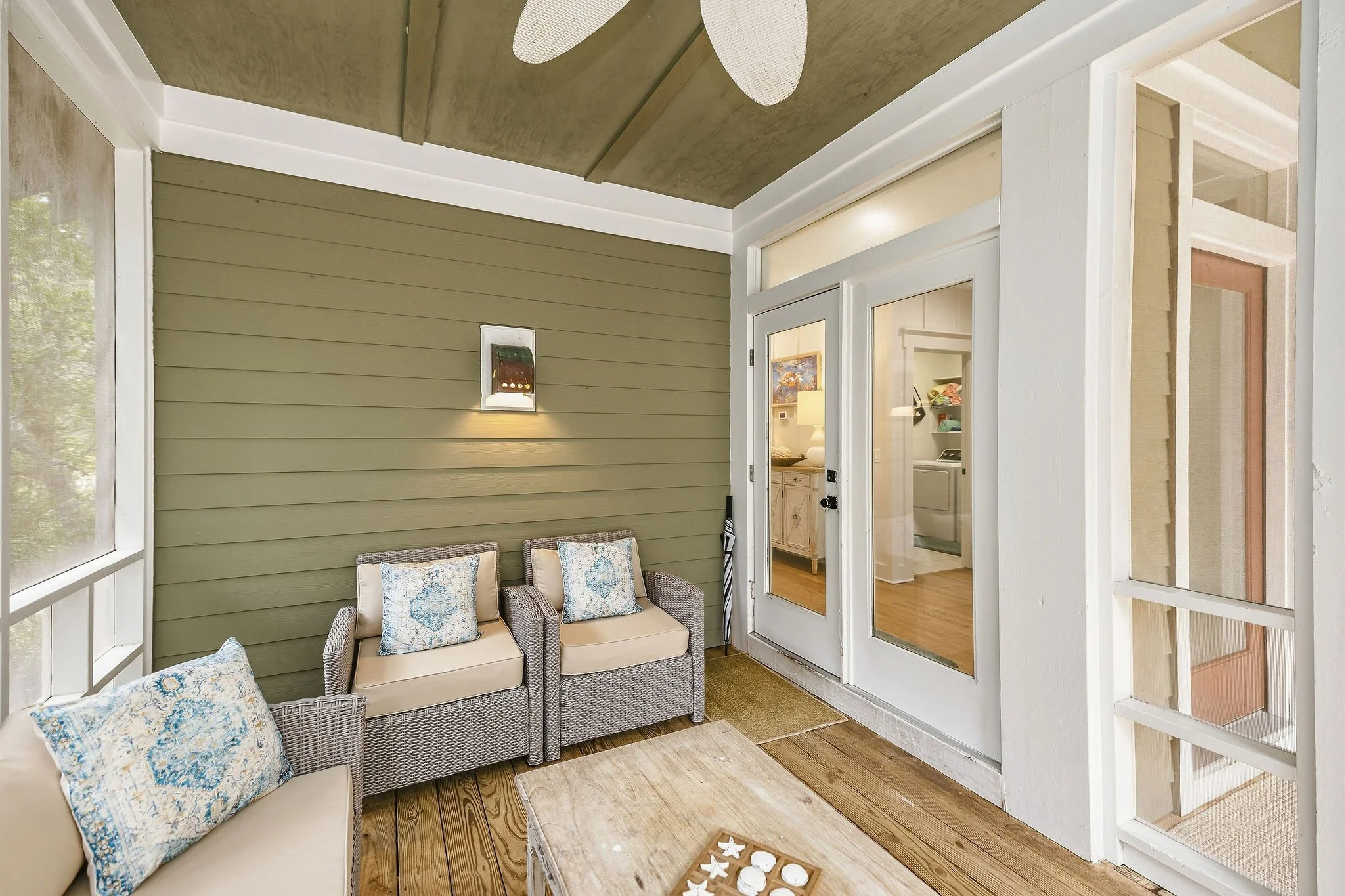 Enclosed porch area with wicker seating and beige cushions, decorative pillows, wooden flooring, and view into the interior of a house with kitchen and living space.