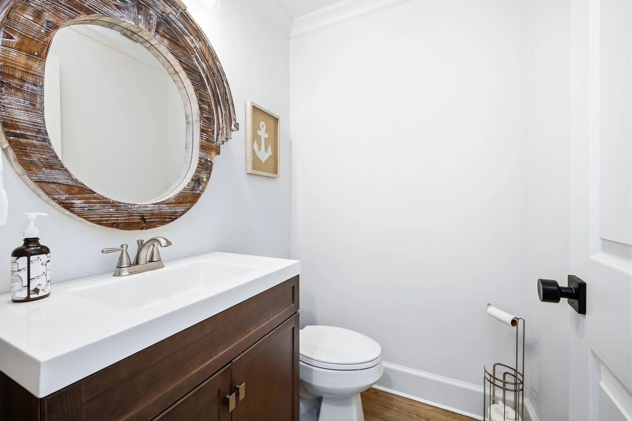 A modern bathroom with a white sink, dark wood vanity, and a round mirror with a rustic wooden frame. There is a framed nautical-themed artwork, a soap dispenser, a toilet, and a black toilet paper holder.