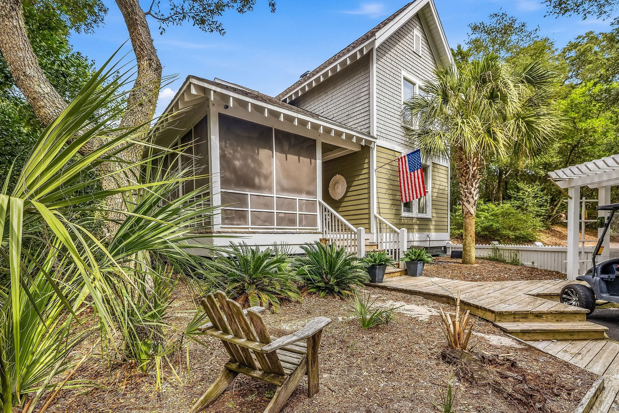 Front yard of a house surrounded by greenery, trees, and plants, with a porch, American flag, and a small walkway.