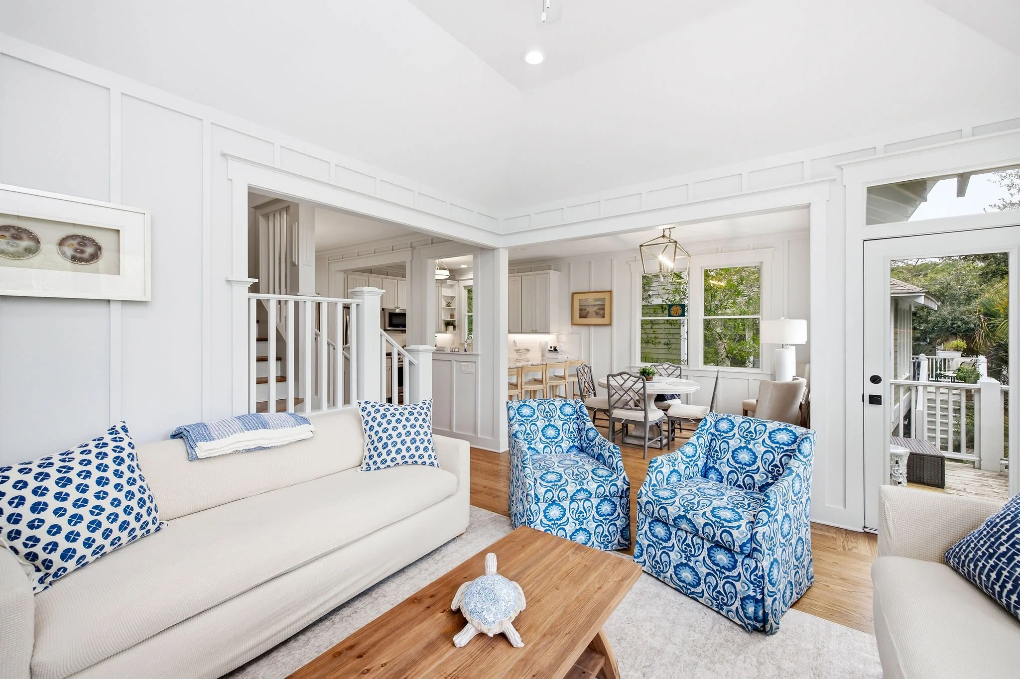 Bright living room with white walls and furniture, patterned blue and white armchairs, beige sofa with blue patterned pillows, wooden coffee table, decorative shell centerpiece, large windows showing trees outside, open kitchen area in the background