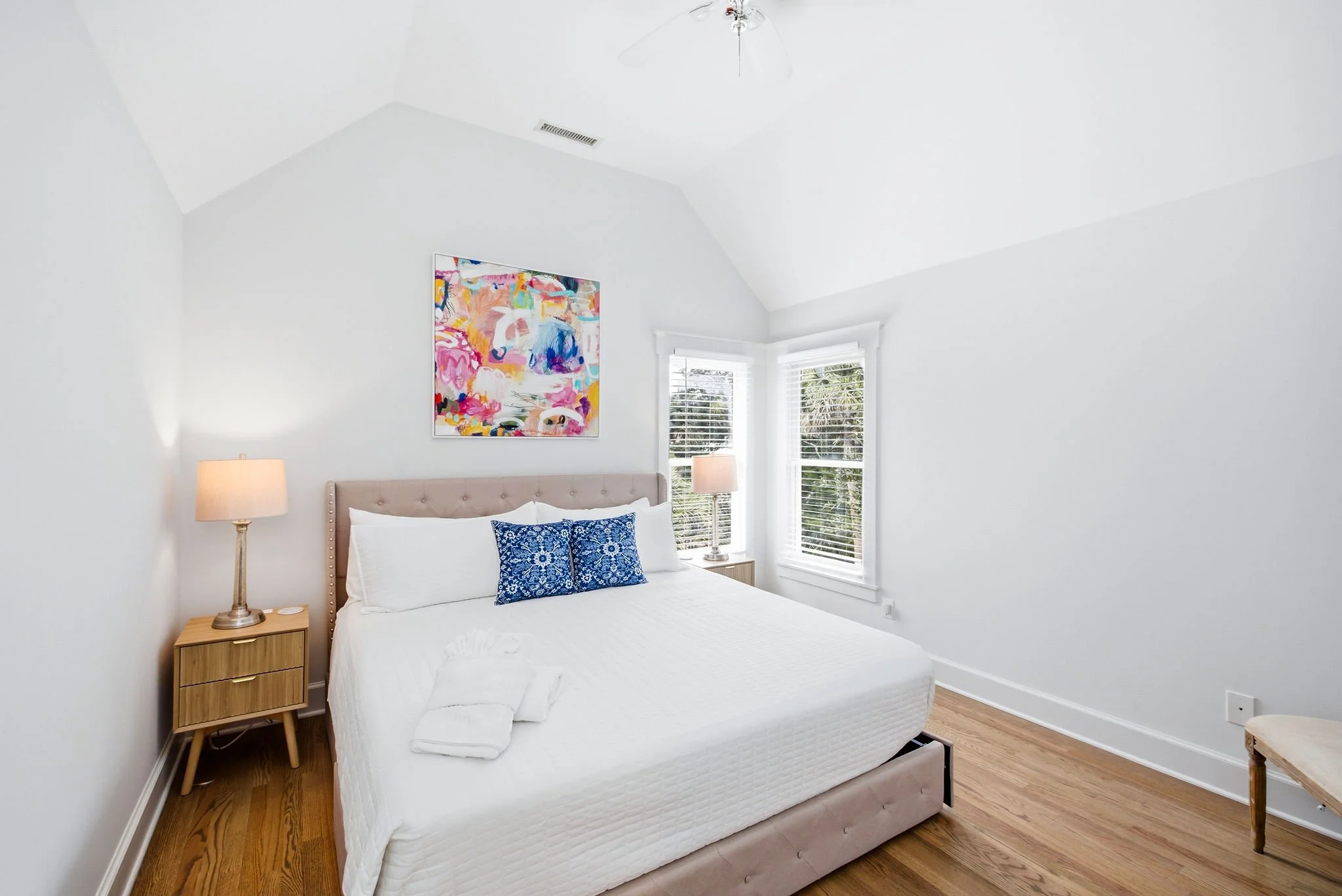 A bright bedroom with a bed, nightstands, lamps, and colorful artwork on the wall.