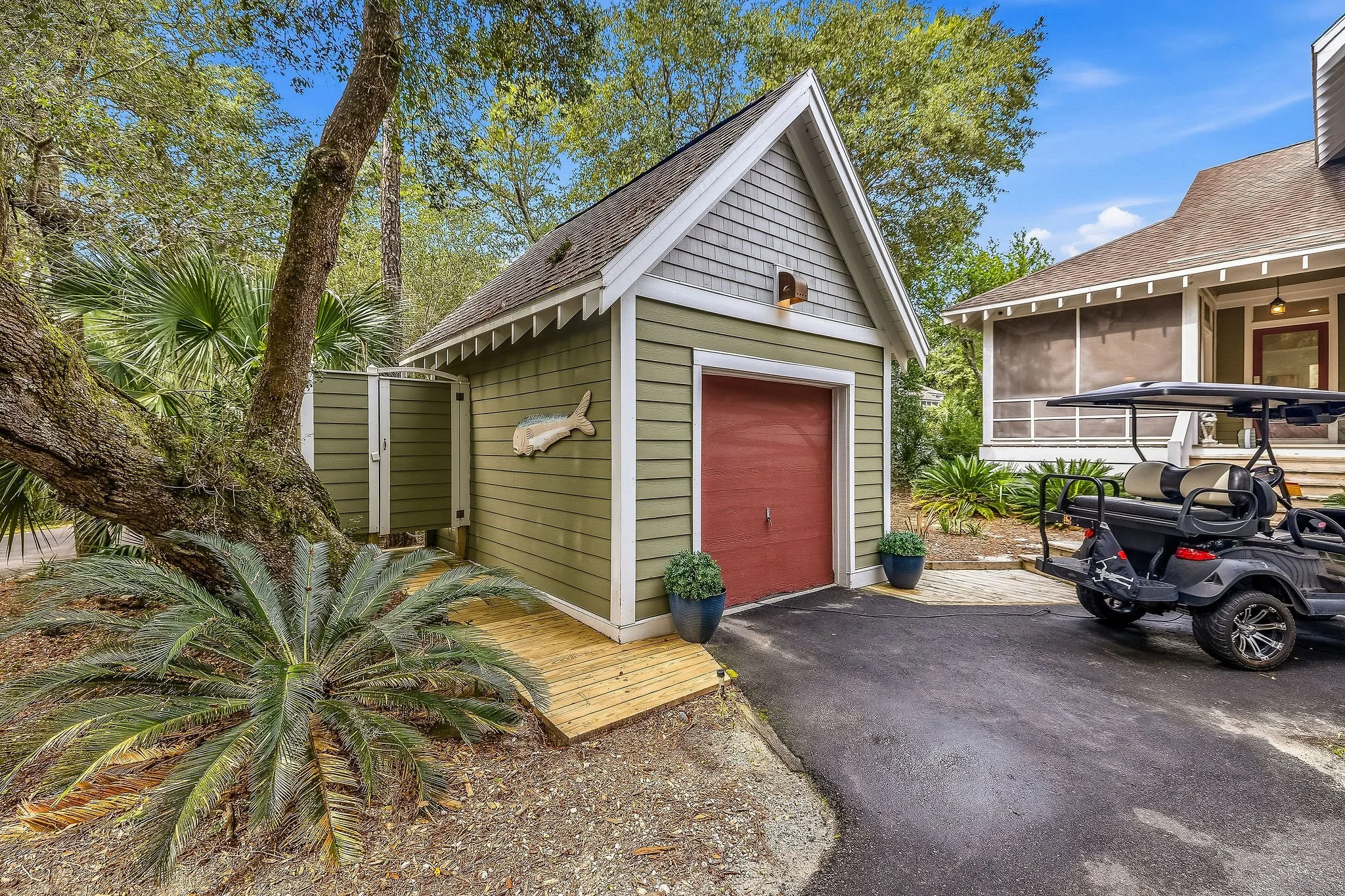 A small green wooden shed with a red door, a fish decoration, two potted plants, surrounded by trees and foliage, with a driveway and a golf cart nearby, part of a house with a screened porch, under a blue sky.