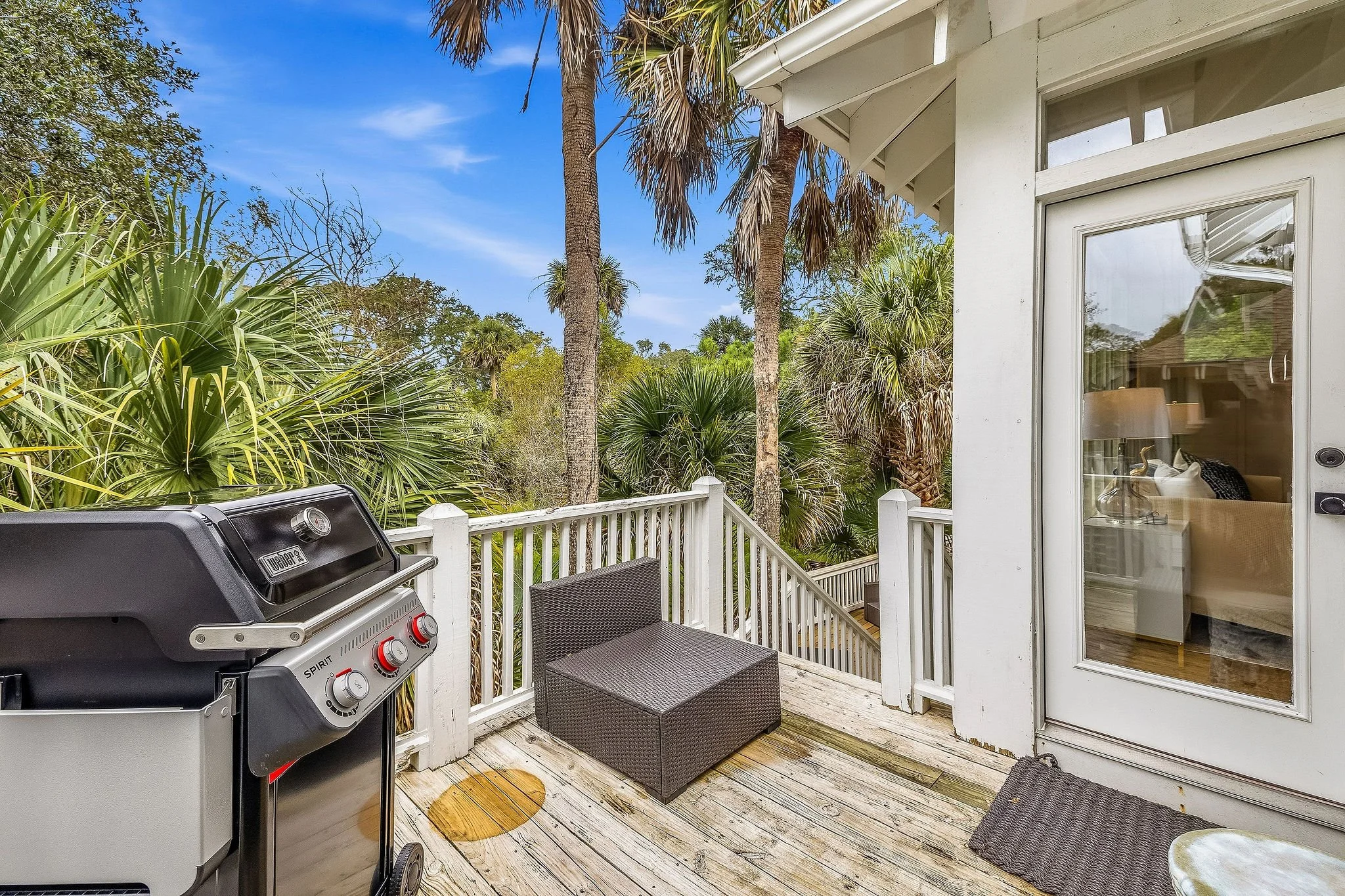 A wooden deck with a gas grill, a wicker chair, and a small rectangular ottoman. The deck is beside a house with a sliding glass door, and overlooks a lush yard with palm trees and greenery under a blue sky.