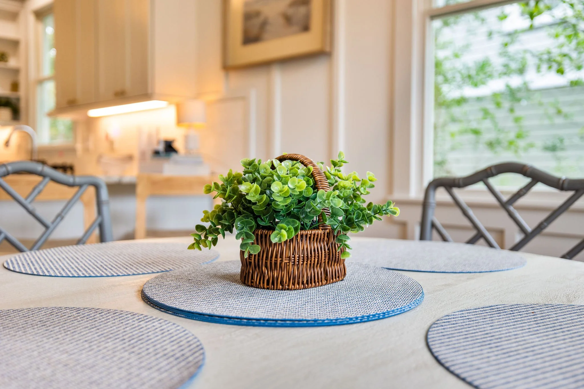 A wicker basket with green artificial plants sits on a dining table with blue and white placemats, in a bright kitchen with large windows and beige walls.