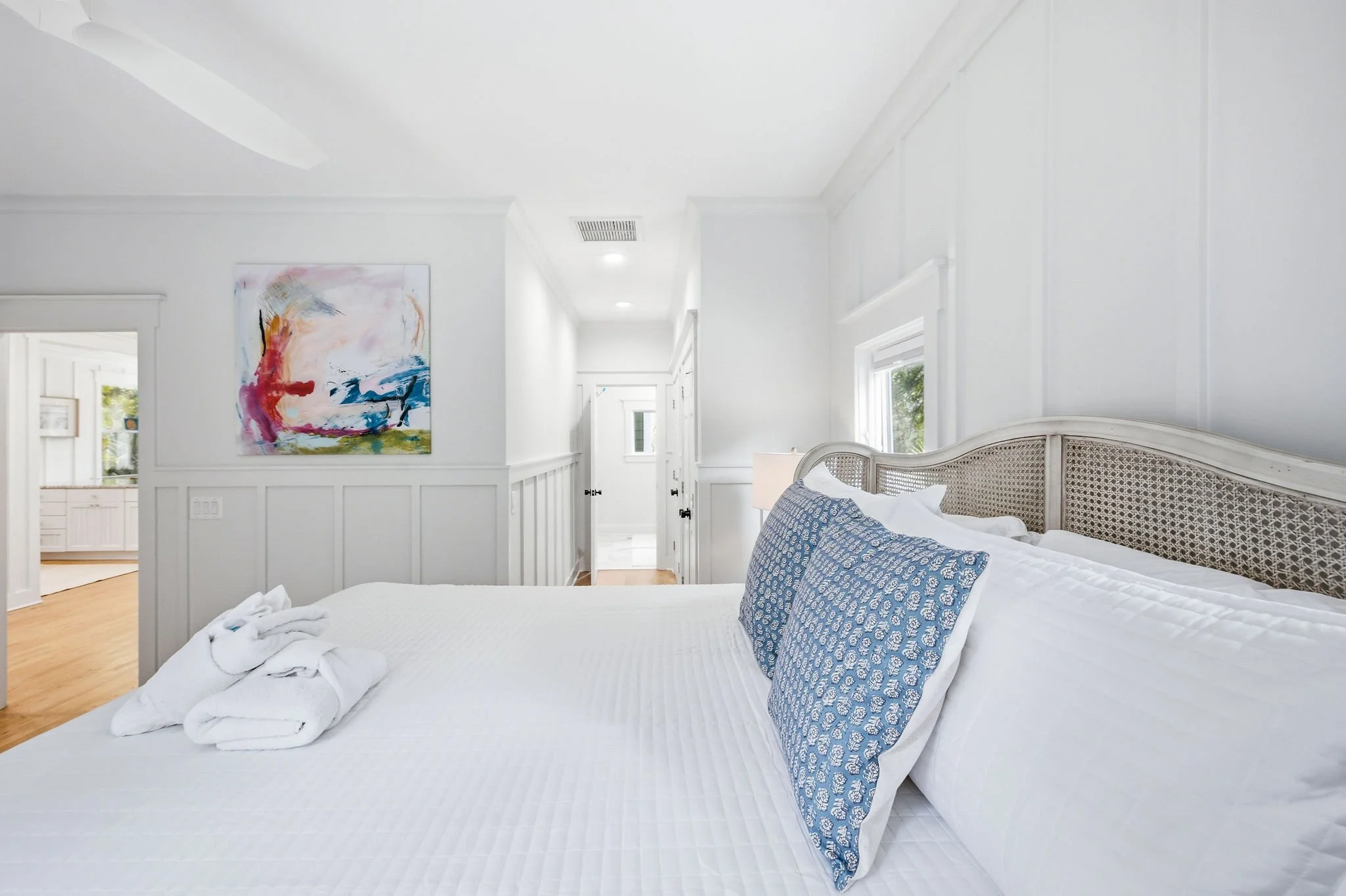 A bright, white bedroom with a white bed that has blue patterned pillows. There is a towel sculpture on the bed, and colorful abstract artwork on the wall. Large windows let in natural light.