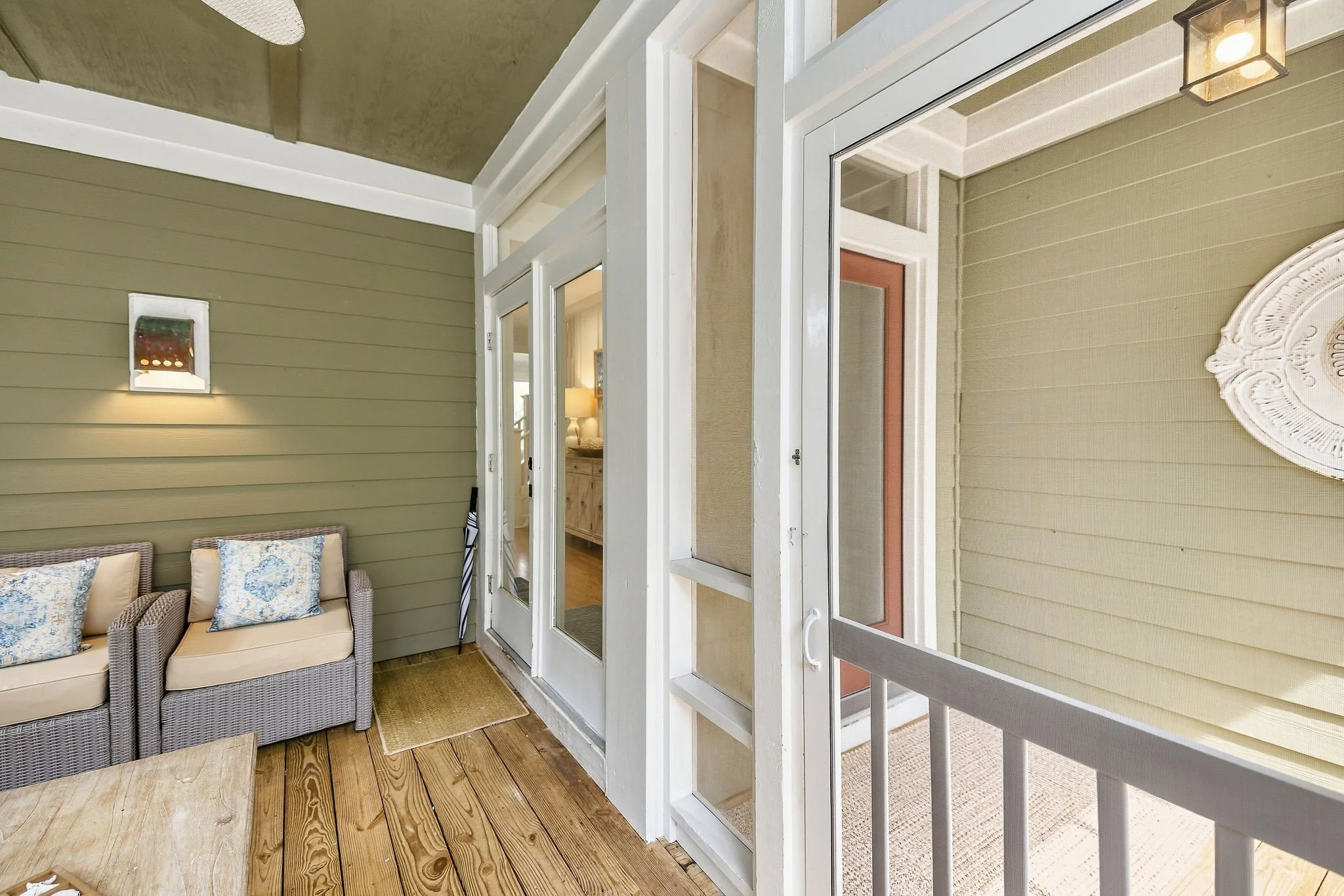 The image shows a covered porch area with a beige porch swing with three pillows, an olive green wall with a wall-mounted light fixture, a wooden floor, and a view of the interior home through a glass door and windows.