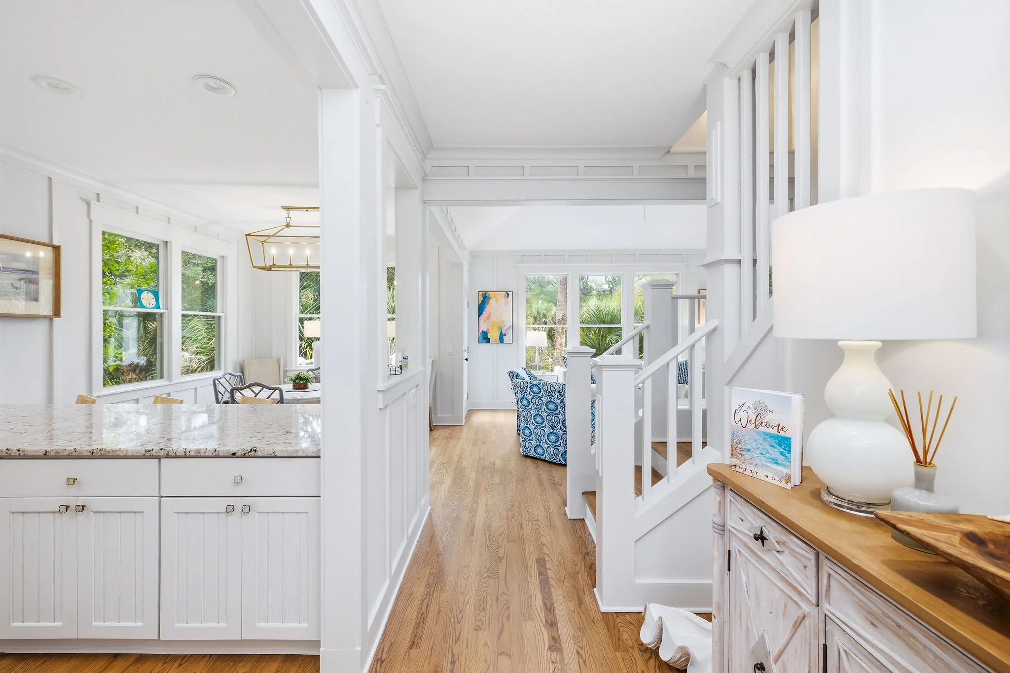 Interior of a bright, white painted living space with large windows showing greenery outside. Features include a kitchen island with a granite countertop, a chandelier light, a sitting area with patterned chairs, and decorative items like lamps, fram