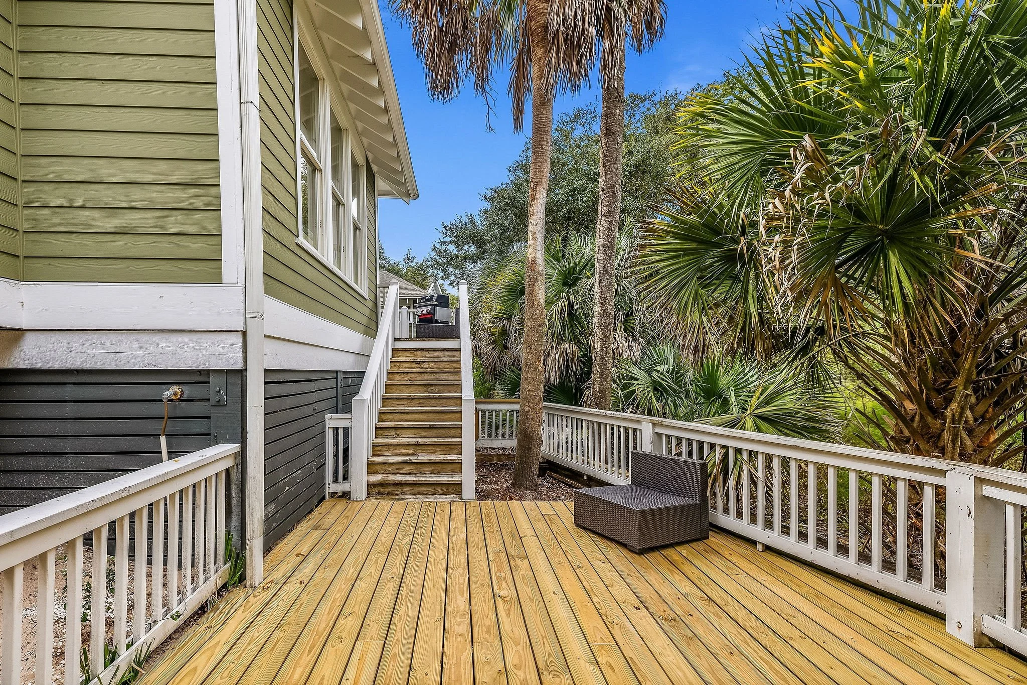 A backyard patio with wooden decking, white railing, a black outdoor chair, and tall palm trees with a clear blue sky in the background.