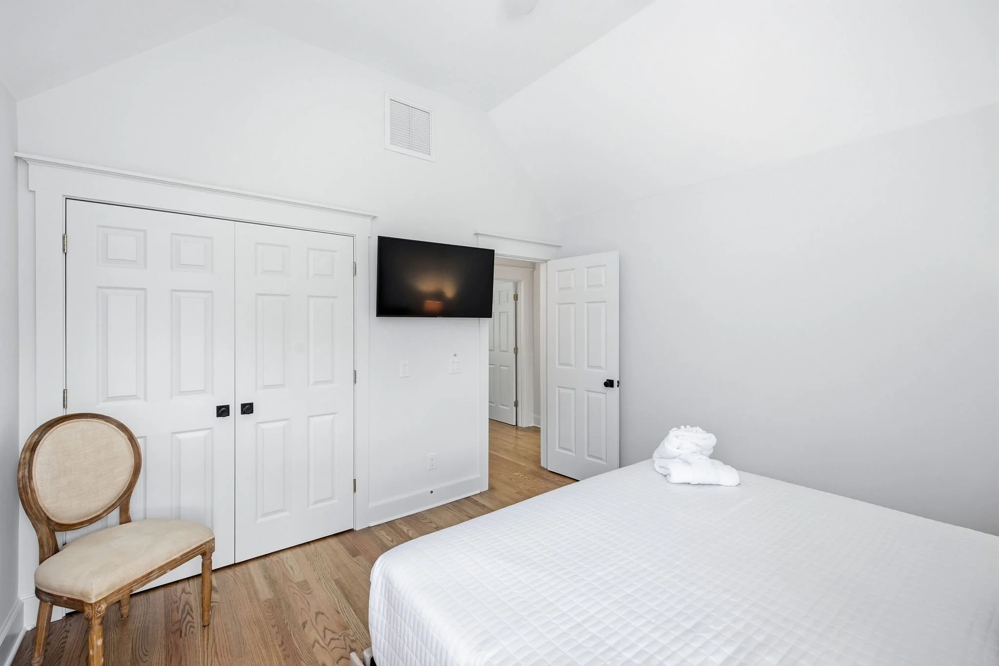 A minimalist bedroom with white walls, a flat-screen TV mounted on the wall, a white bed with folded white towels on it, a beige upholstered chair, and a closet with white double doors.