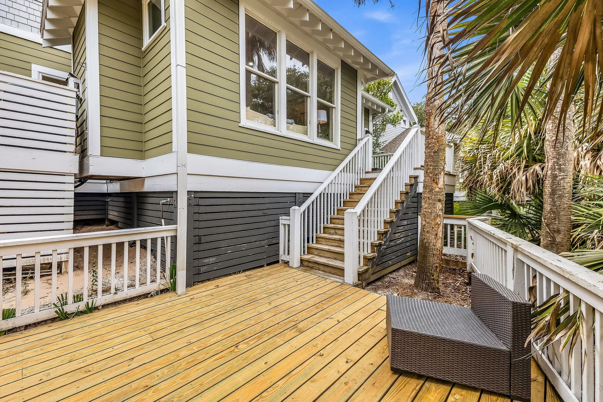 Backyard deck with wooden flooring, white railings, stairs leading up to a house with green siding, large window, and palm trees on the side.