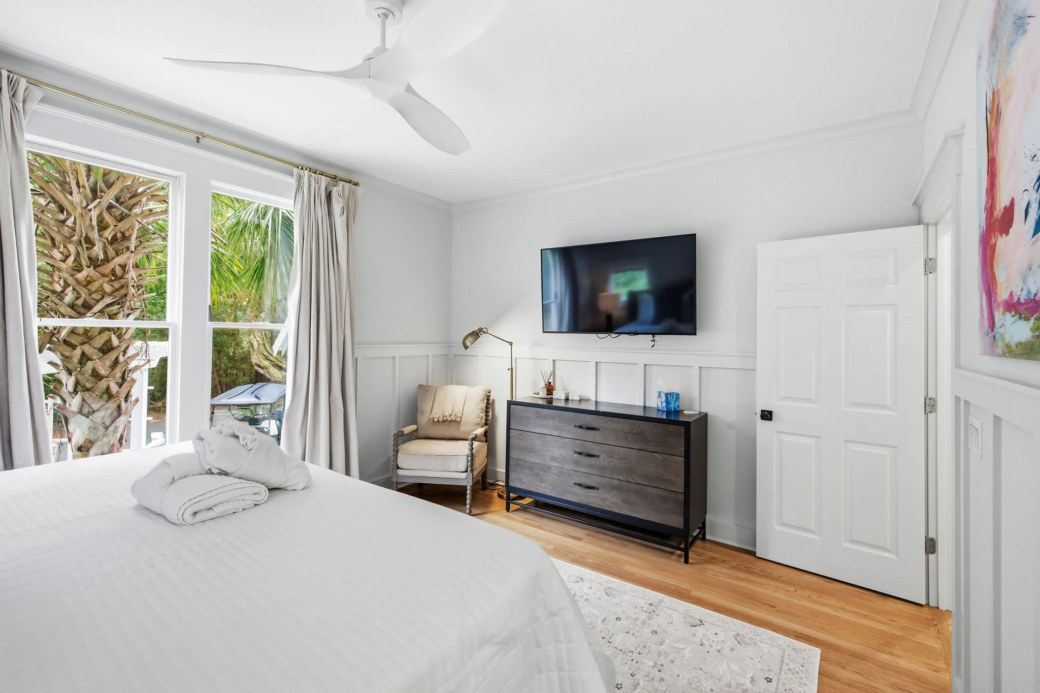 Bright bedroom with white walls, large windows with cream curtains showing greenery outside, a bed with folded towels, a beige armchair, a wooden dresser, a wall-mounted flat-screen TV, a silver floor lamp, and a colorful abstract painting.