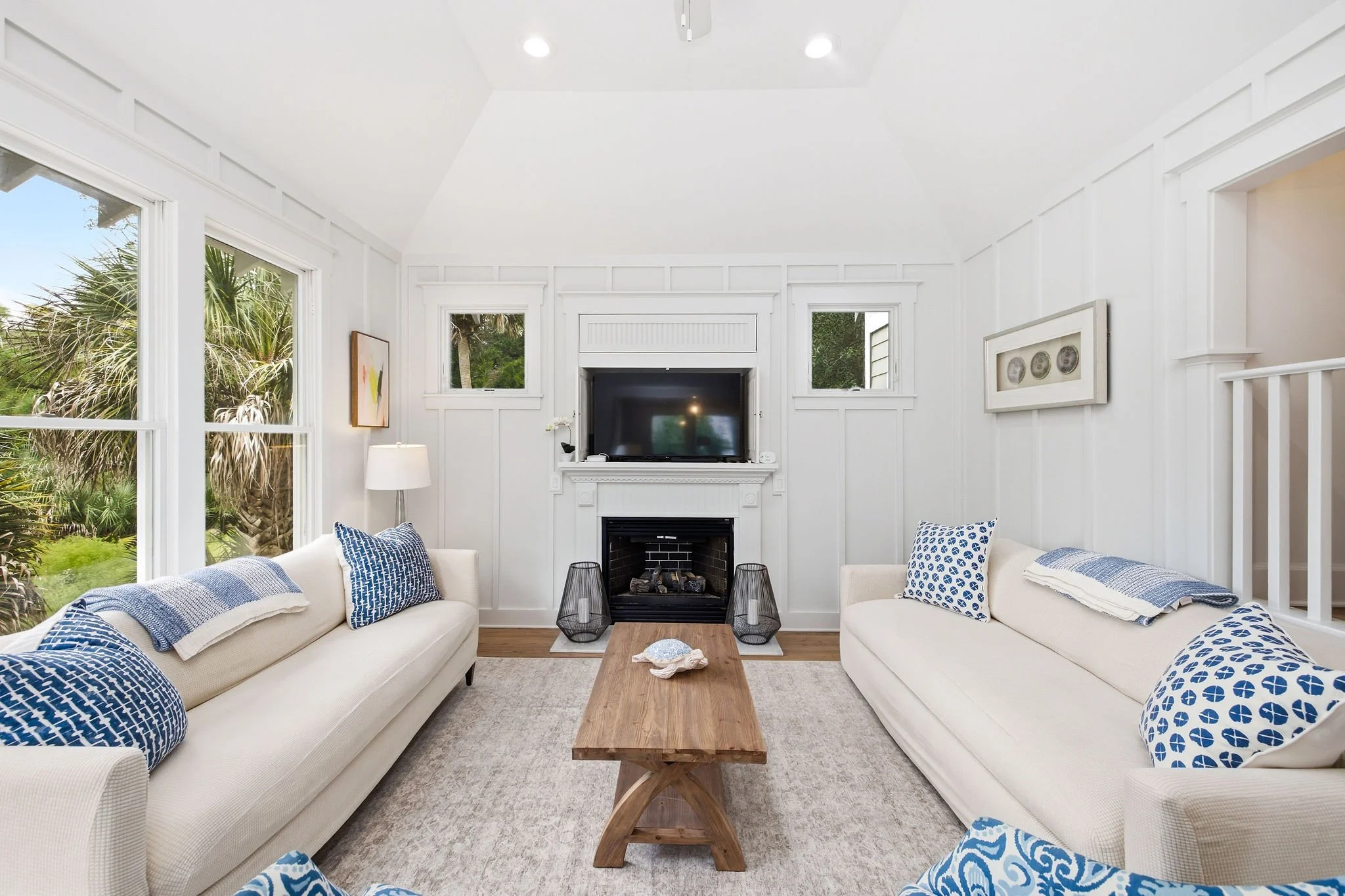 A cozy living room with white walls and trim, two white sofas with blue patterned cushions, a wooden coffee table, a fireplace, a flat-screen TV above the fireplace, a white table lamp, and large windows showing green outdoor scenery.