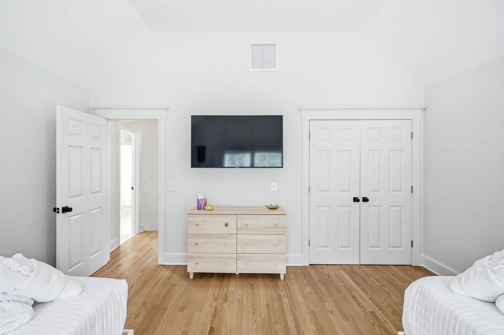 A bedroom with two white beds, a wooden dresser with items on top, and a wall-mounted flat-screen TV. The room has white walls, a ceiling with an air vent, and wooden flooring.