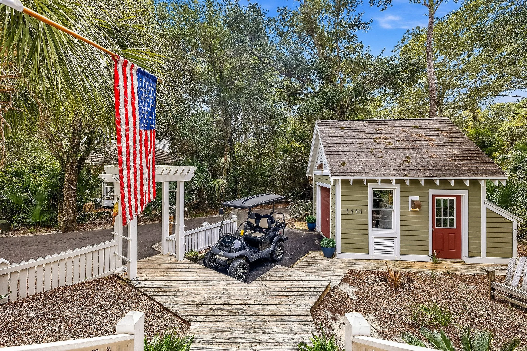 A small green house with a red door, white trim, and a shingled roof is surrounded by trees. An American flag hangs from a pole on the left side of the image. There is a wooden deck and a paved driveway with a golf cart parked on it. A white picket f