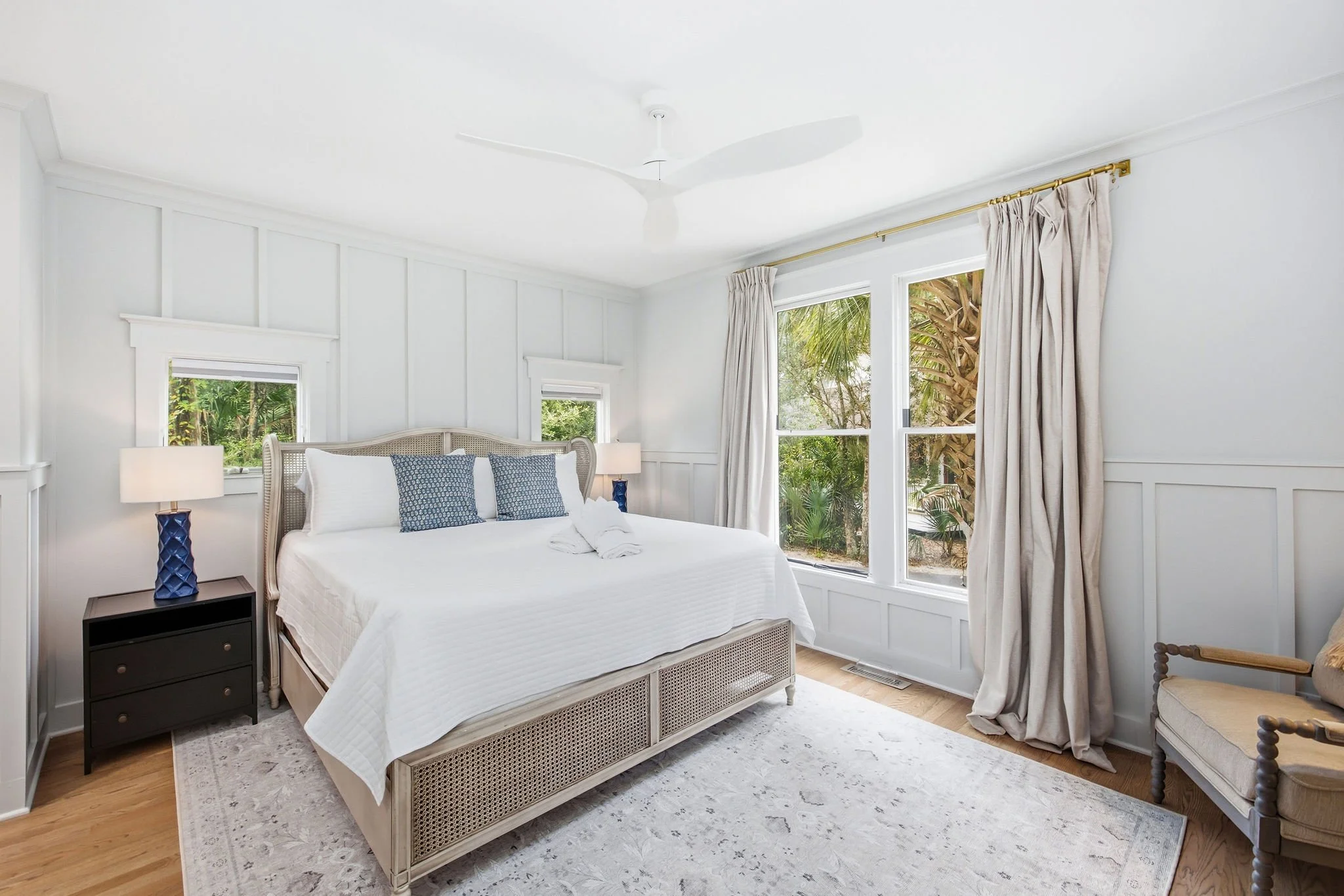 Bright bedroom with white walls, large windows with curtains showing a garden view, a white bed with blue and white pillows, black bedside table with a blue lamp, and a ceiling fan.