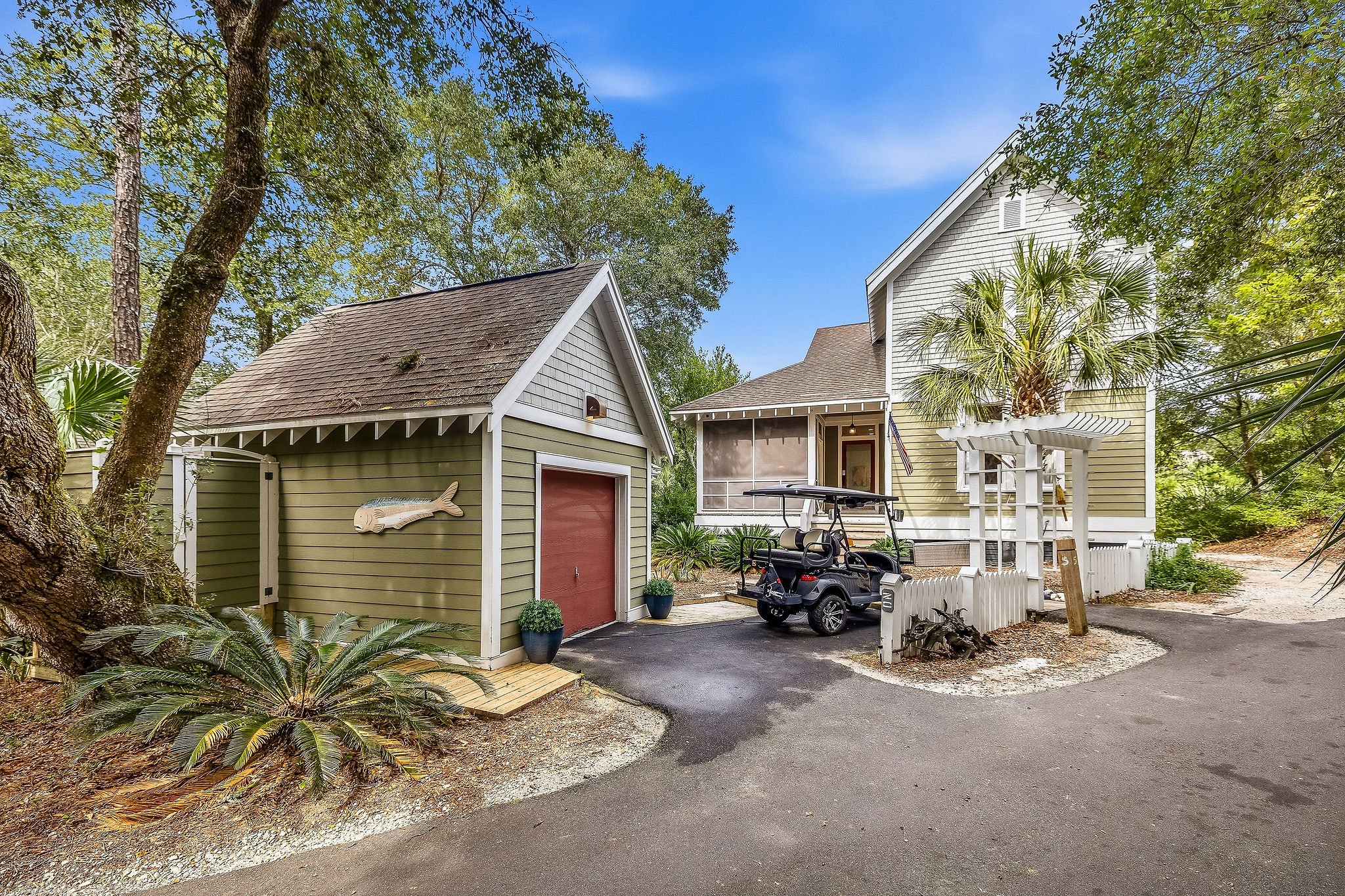 A driveway leading to a house with a small roofed porch, surrounded by trees and greenery under a blue sky with some clouds. There is a golf cart parked on the driveway and a small shed with a fish decoration on the side.