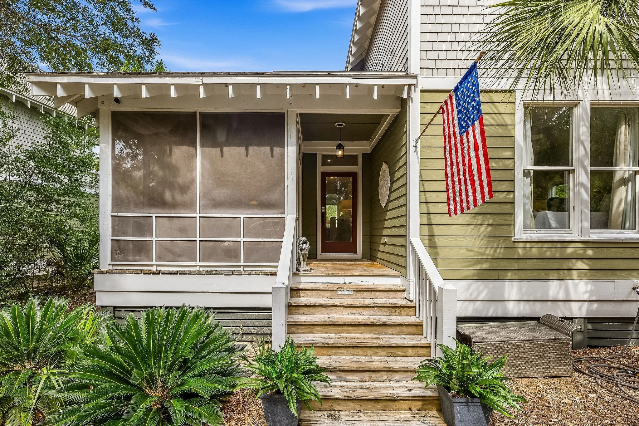 The image shows the front porch of a house with wooden stairs leading up to a door. There is an American flag hanging on the front of the house. The house has beige and white siding, and there are trees and plants around the porch, with a clear blue 