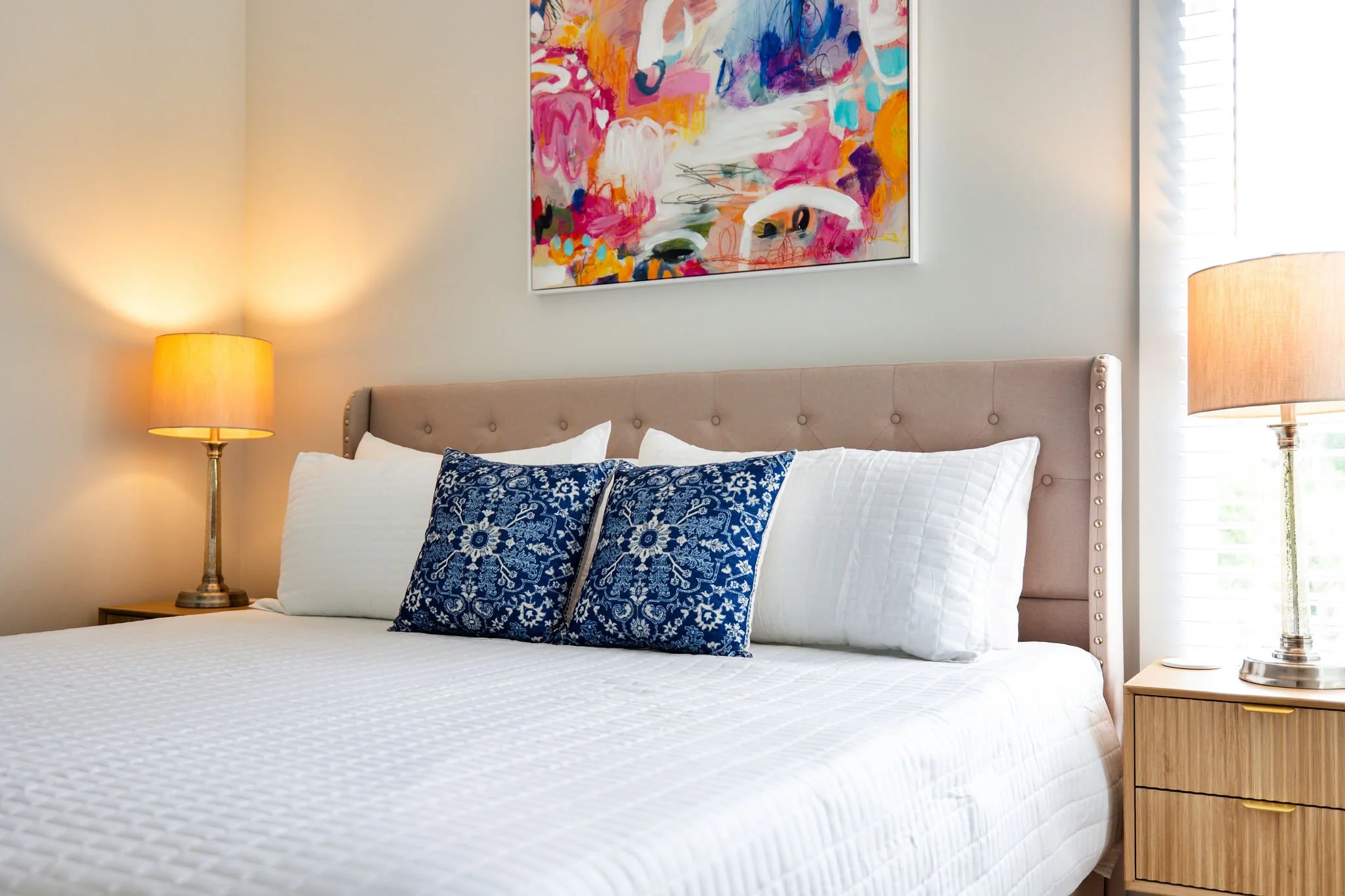 A bedroom with a beige upholstered bed headboard, white bedding, two blue patterned throw pillows, two side tables with lamps, a colorful abstract painting on the wall, and window blinds letting in natural light.