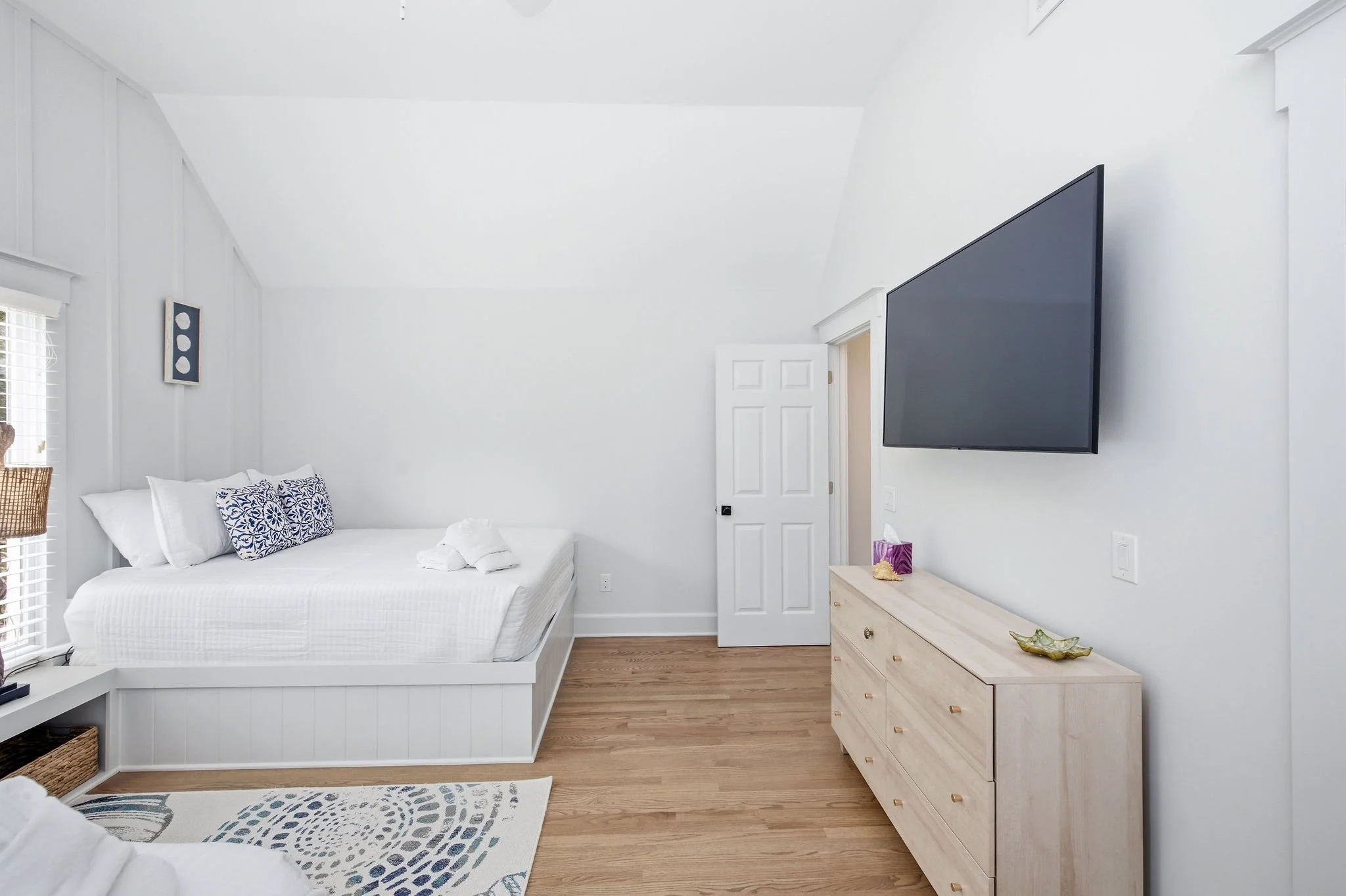 A bright bedroom with white walls, a white bed with decorative pillows, a wooden dresser with jewelry on top, a large wall-mounted TV, and a window with blinds.
