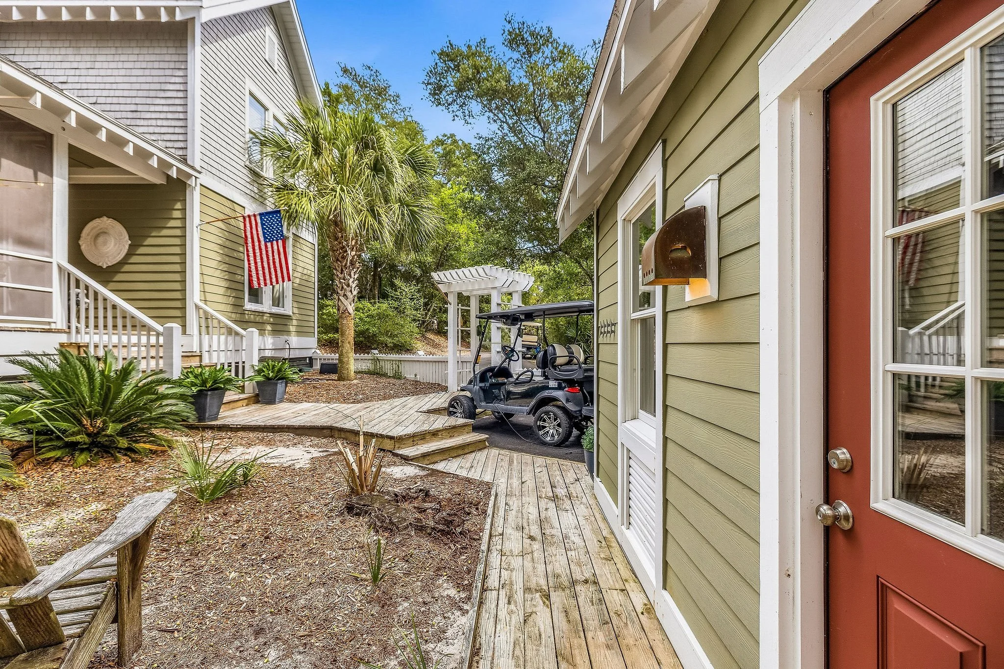 A backyard patio area with a wooden deck, green and beige sidelighted houses, palm trees, bushes, a golf cart, and an American flag hanging on a house wall.