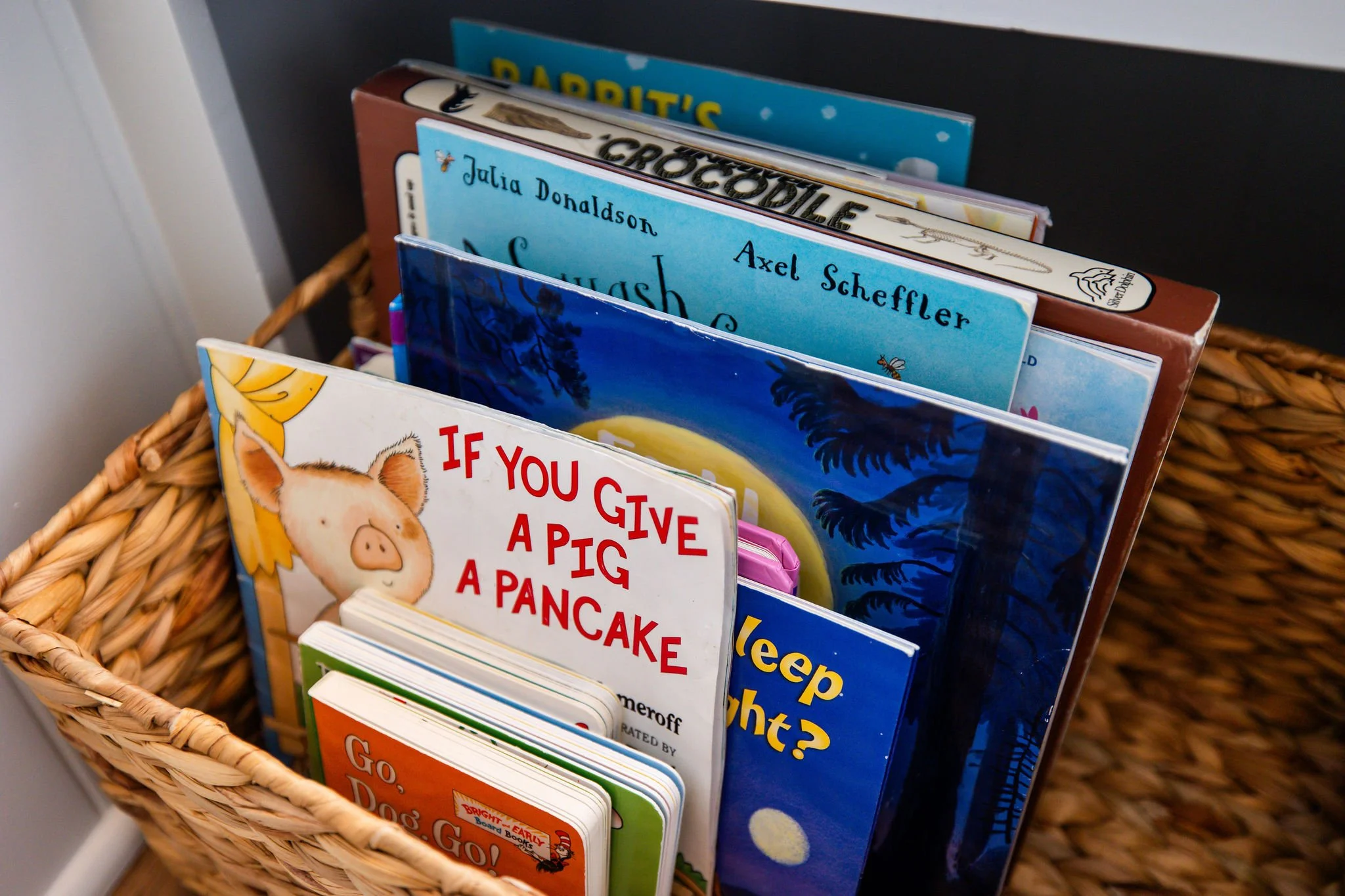 A wicker basket holding children's books in an upright position, including titles like 'If You Give a Pig a Pancake' and others, with a black background.