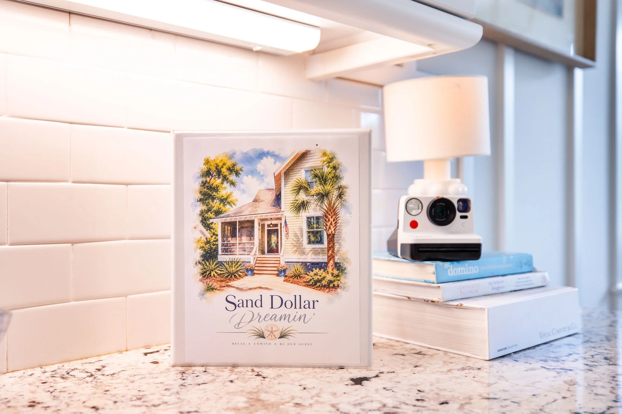 A countertop with a colorful brochure featuring a house, with the title 'Sand Dollar Dreamin',' and a small white camera and several books stacked beside it.