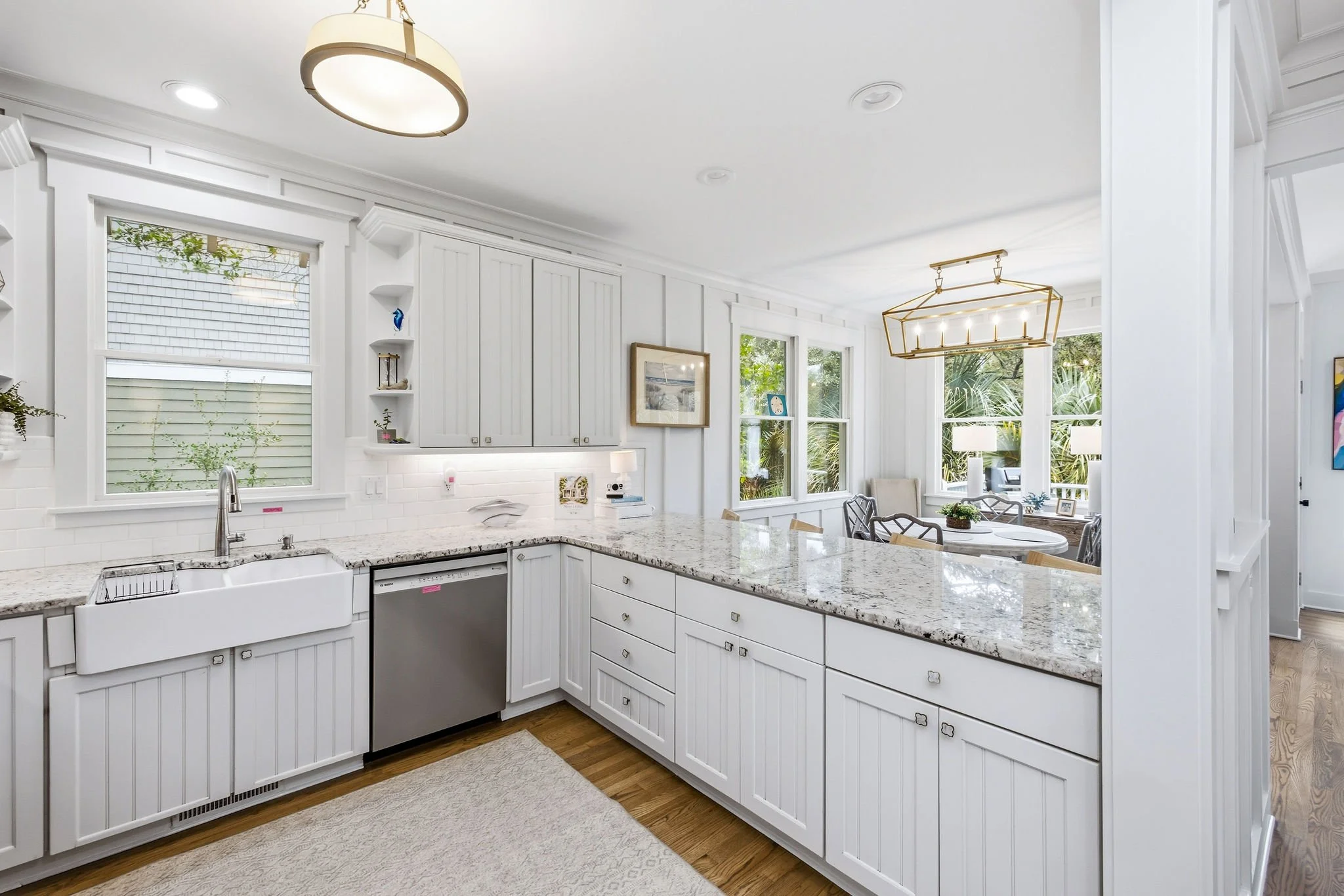 Bright modern kitchen with white cabinets, granite countertops, stainless steel dishwasher, white farmhouse sink, large windows with greenery outside, and a dining area with a gold chandelier.