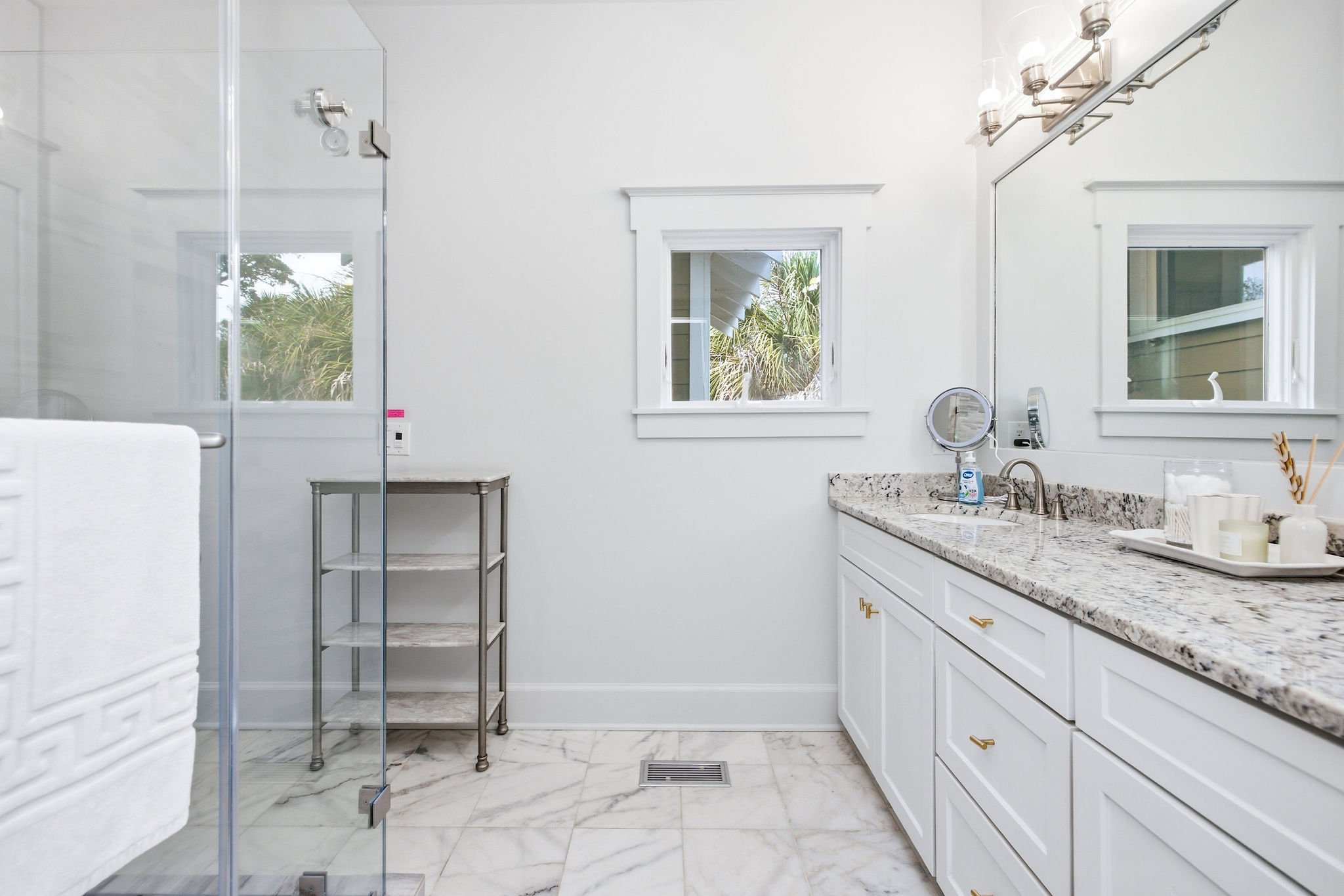 A bright bathroom with white walls, a double vanity with granite countertop, two windows showing green foliage outside, and a glass shower enclosure on the left.