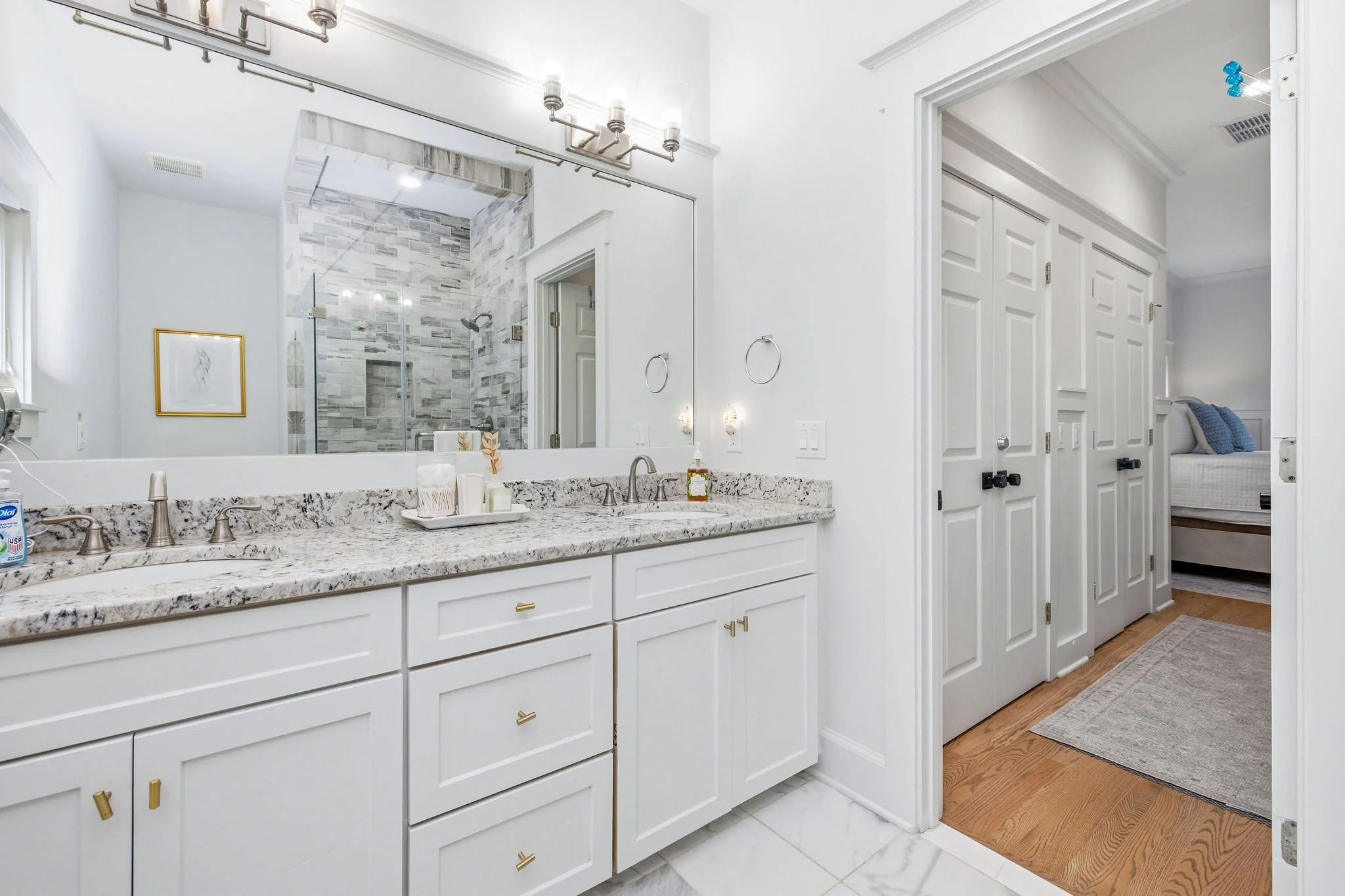 Bathroom with white cabinets, granite countertop, large mirror, and a ceiling light fixture. A shower with gray tile walls and a glass door is visible in the reflection, and a bedroom with a bed and blue pillows is seen through an open door.