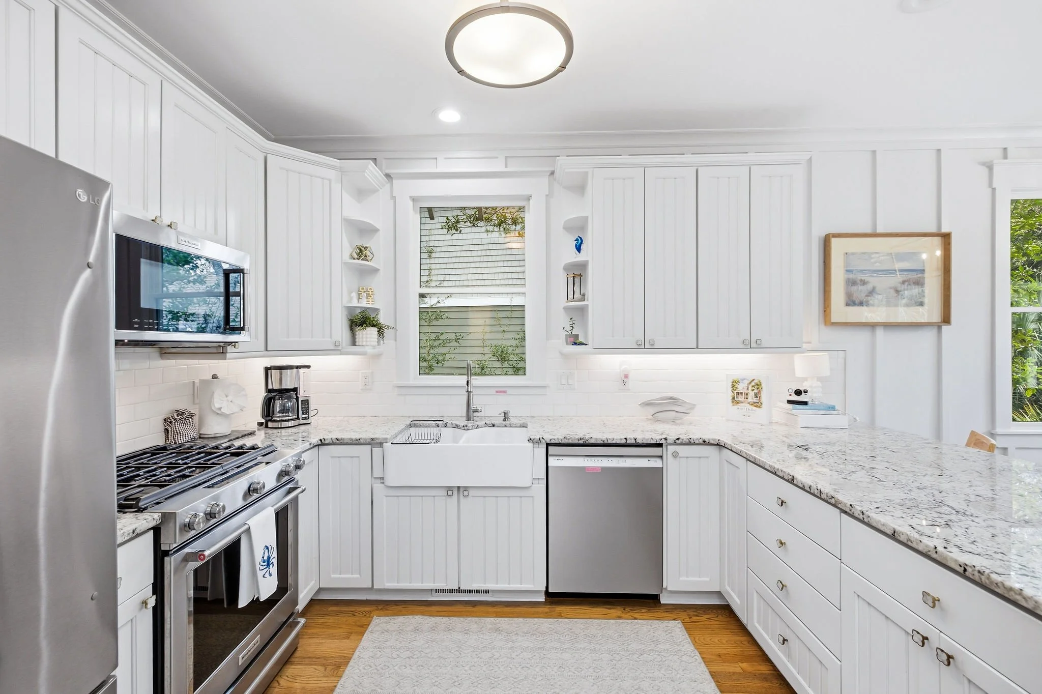 A bright white kitchen with white cabinets, a granite countertop, a stainless steel oven and refrigerator, and a window overlooking greenery.