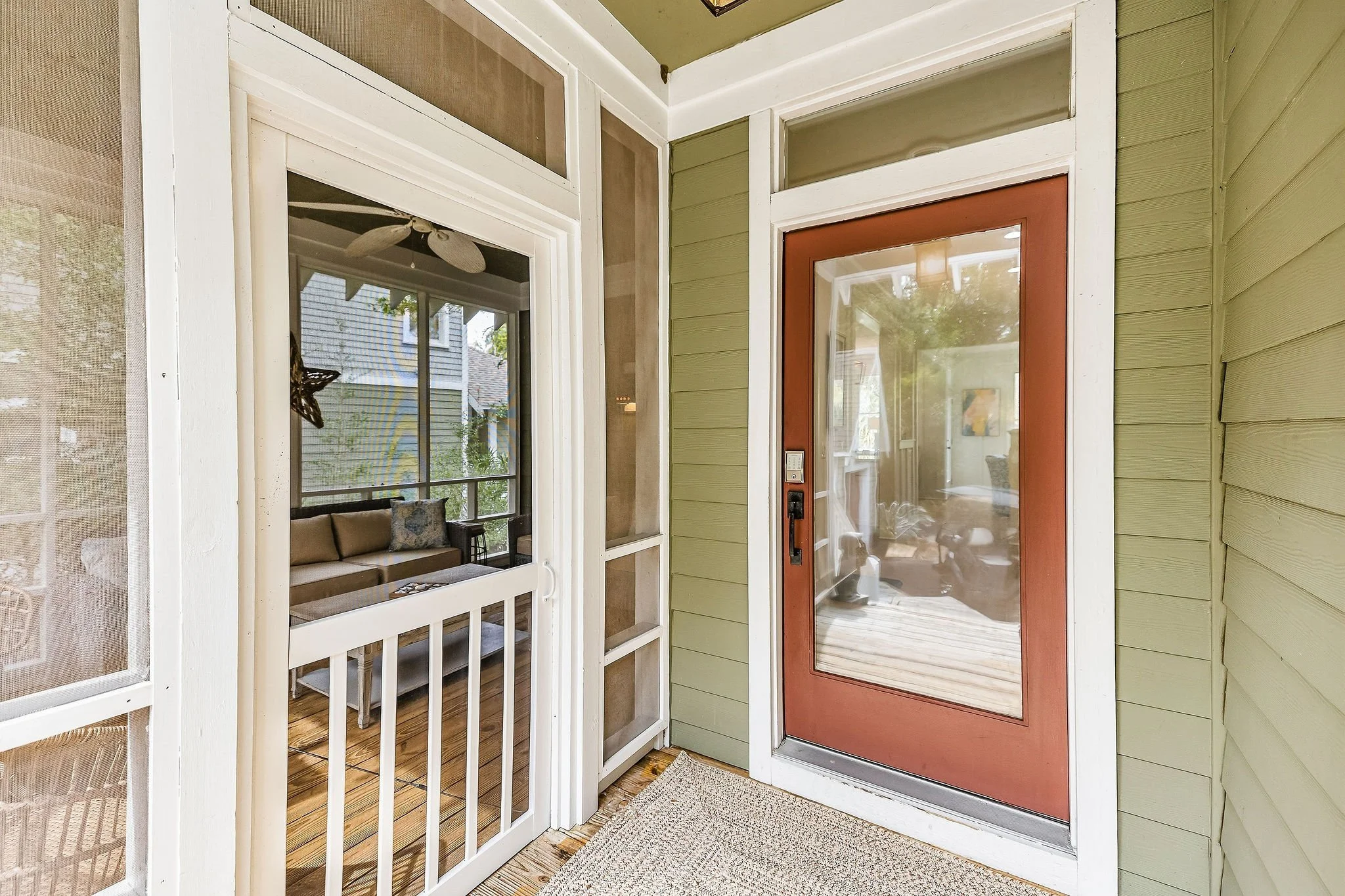 View of front porch area showing red door with glass panel, green siding, a screen door, and partially visible interior with seating and windows.