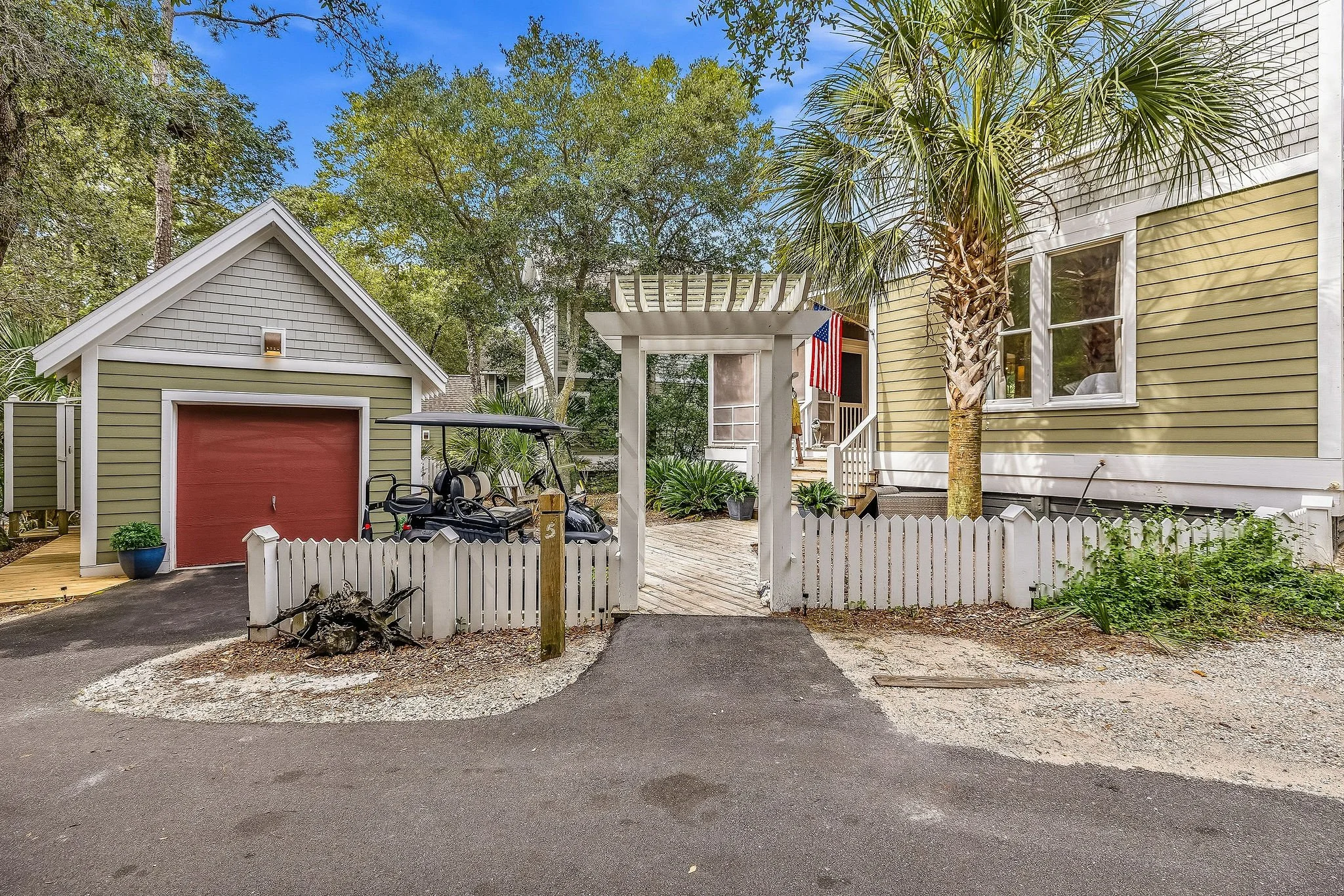 Front view of a yellow house with white trim, a palm tree, a small shed with a red door, a golf cart, and a white picket fence. An American flag hangs near the porch, and there is greenery and trees around.