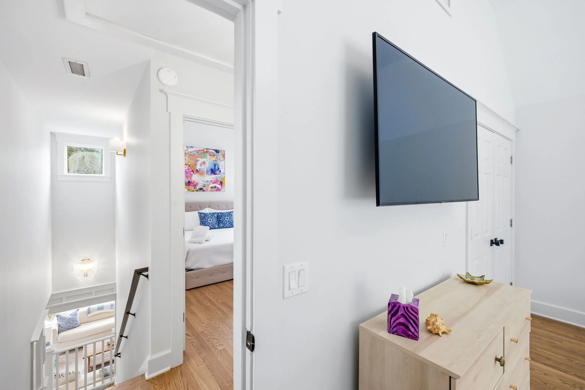 Interior of a house showing a hallway with a living room and bedroom. A flat-screen TV is mounted on the wall. There is a wooden console table with a purple tissue box and a decorative shell. The bedroom has a bed with blue pillows and a colorful pai