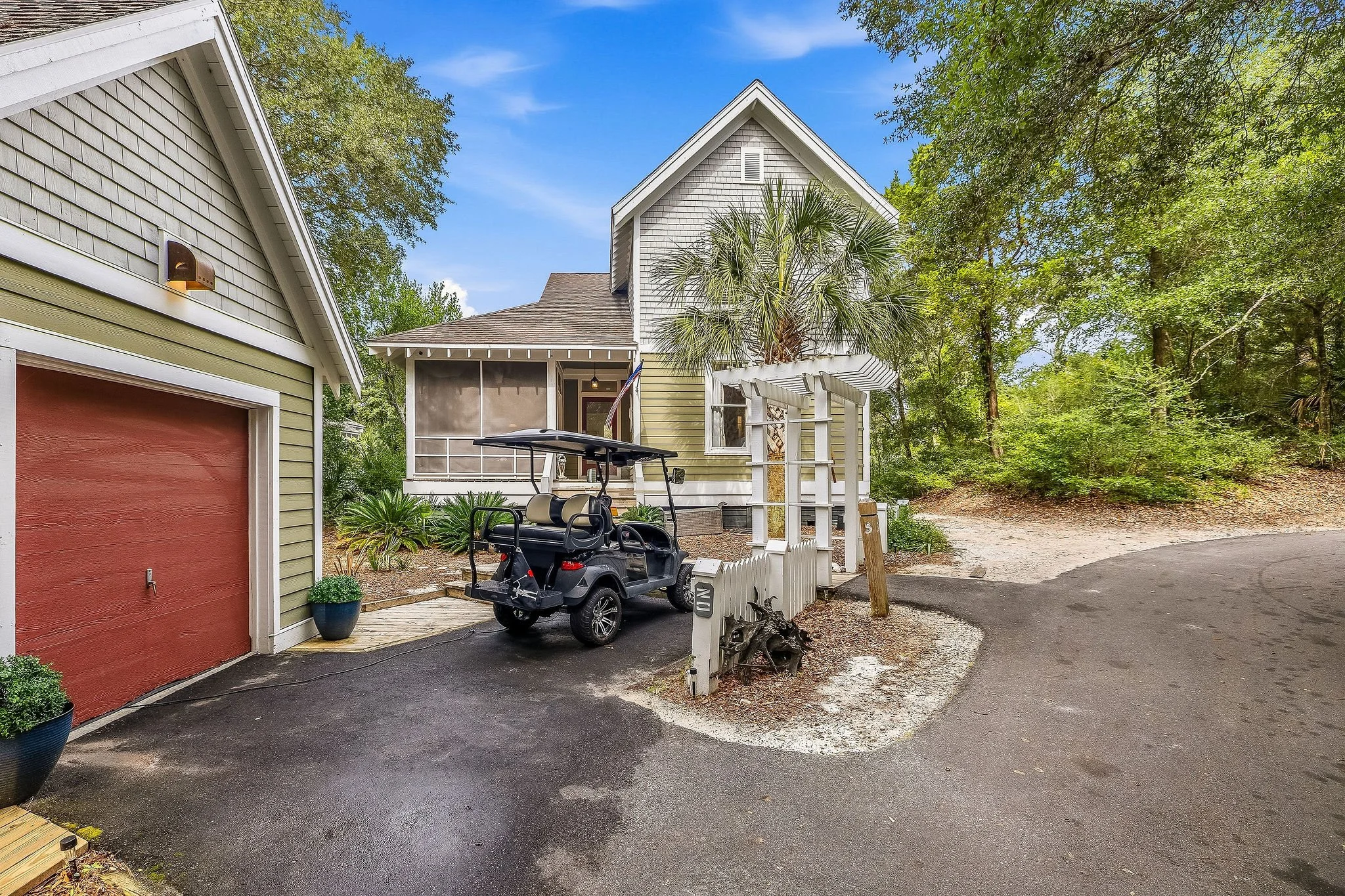 A small house with light yellow siding and a gray roof, surrounded by trees with a clear blue sky above. In front, there is a black golf cart parked on a paved driveway, with some potted plants nearby. A white picket fence is partially visible, and a