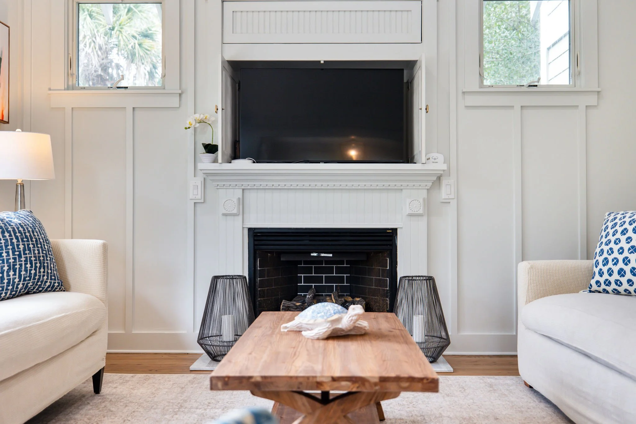 A living room with a fireplace, two white sofas with patterned cushions, a wooden coffee table, and two black wireframe lamps. There are two windows on either side of the fireplace and some decorative items including a potted plant and a tissue box.